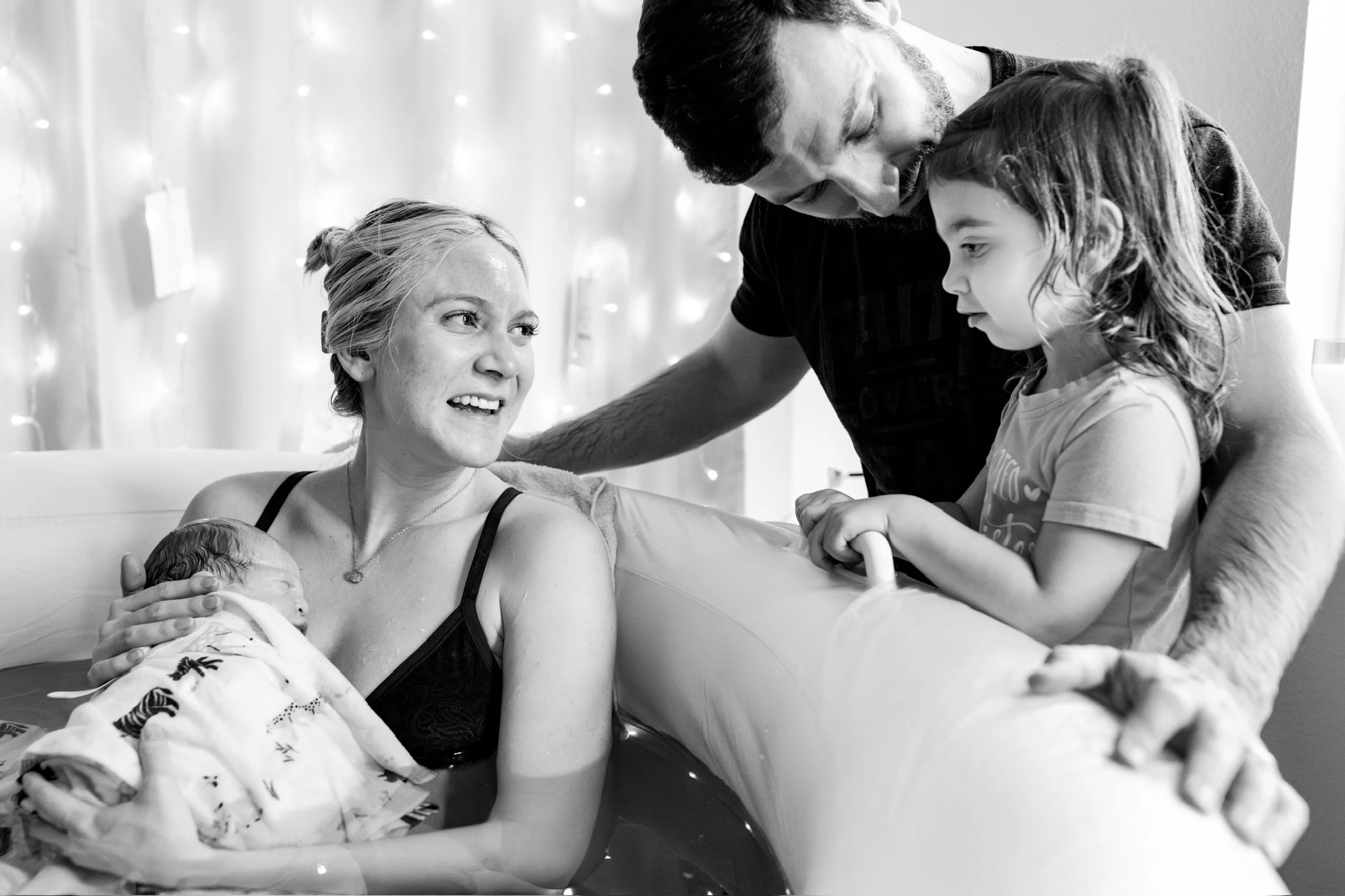 Mother holding newborn baby in birthing pool, with a father and young child nearby, in a black and white photo.