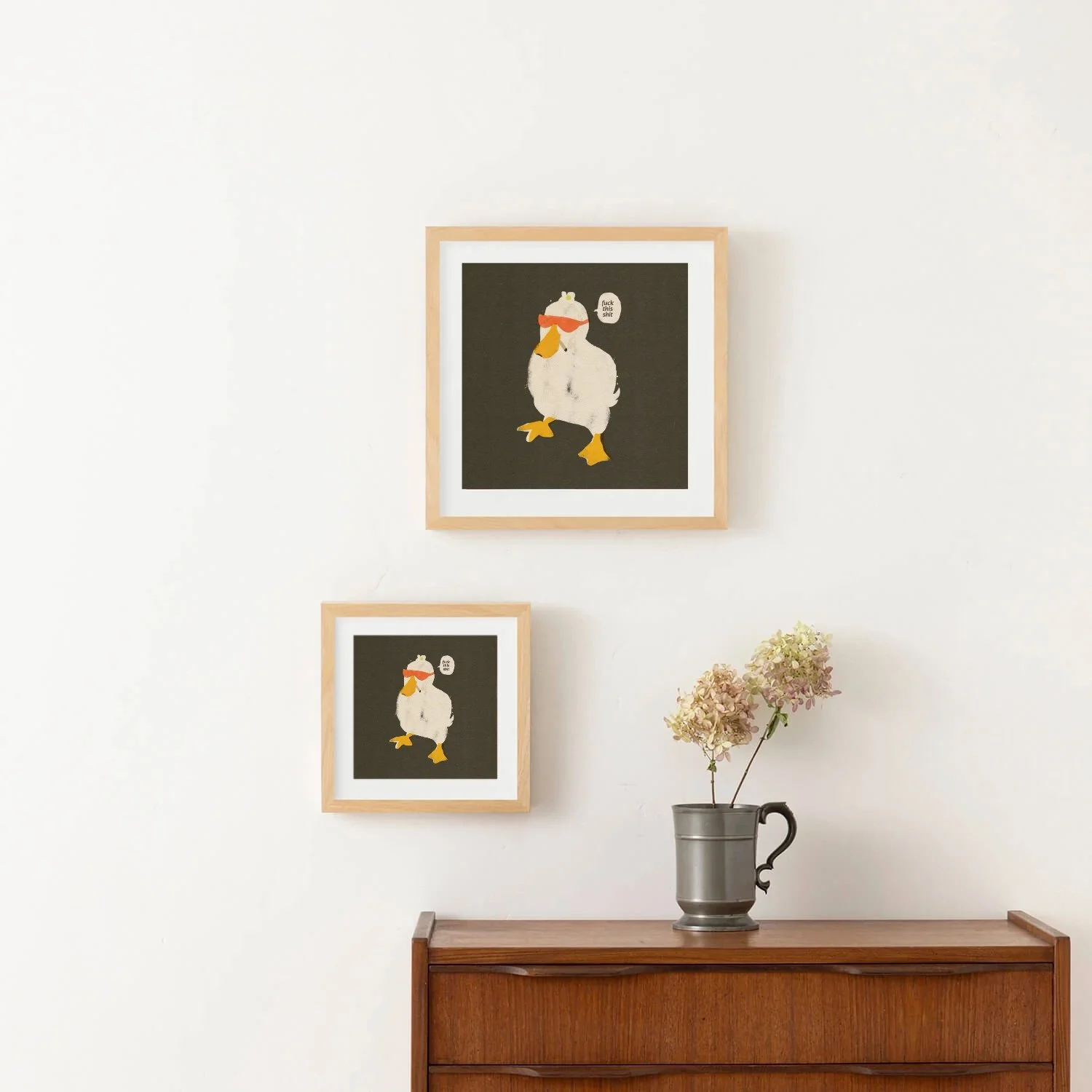 Two framed sarcastic duck art prints hanging on a light wall above a vintage wooden cabinet with dried flowers.