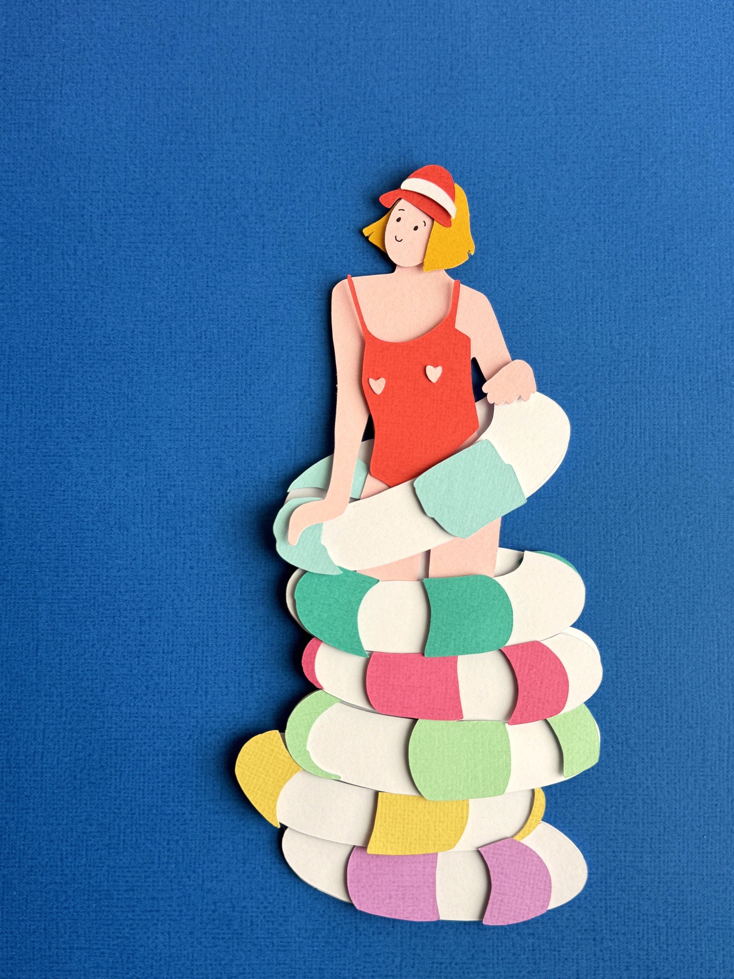 Paper craft of a girl with yellow hair in a red swimsuit and red hat, sitting on colorful pool floats against a blue background.