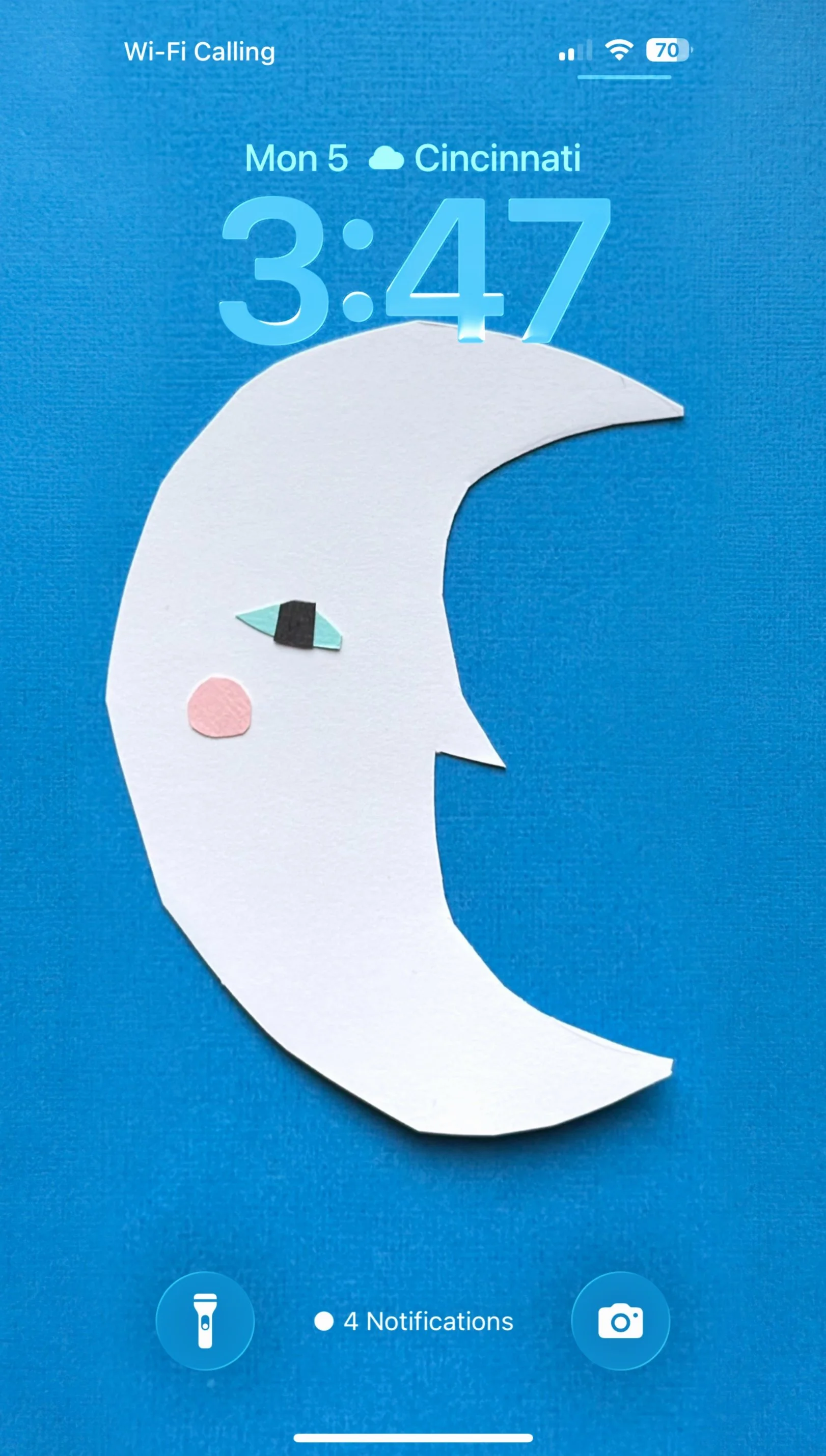 Smartphone lock screen showing time 3:47, date Monday the 5th, weather in Cincinnati, a crescent moon with a face made of geometric paper shapes, Wi-Fi calling, and notifications