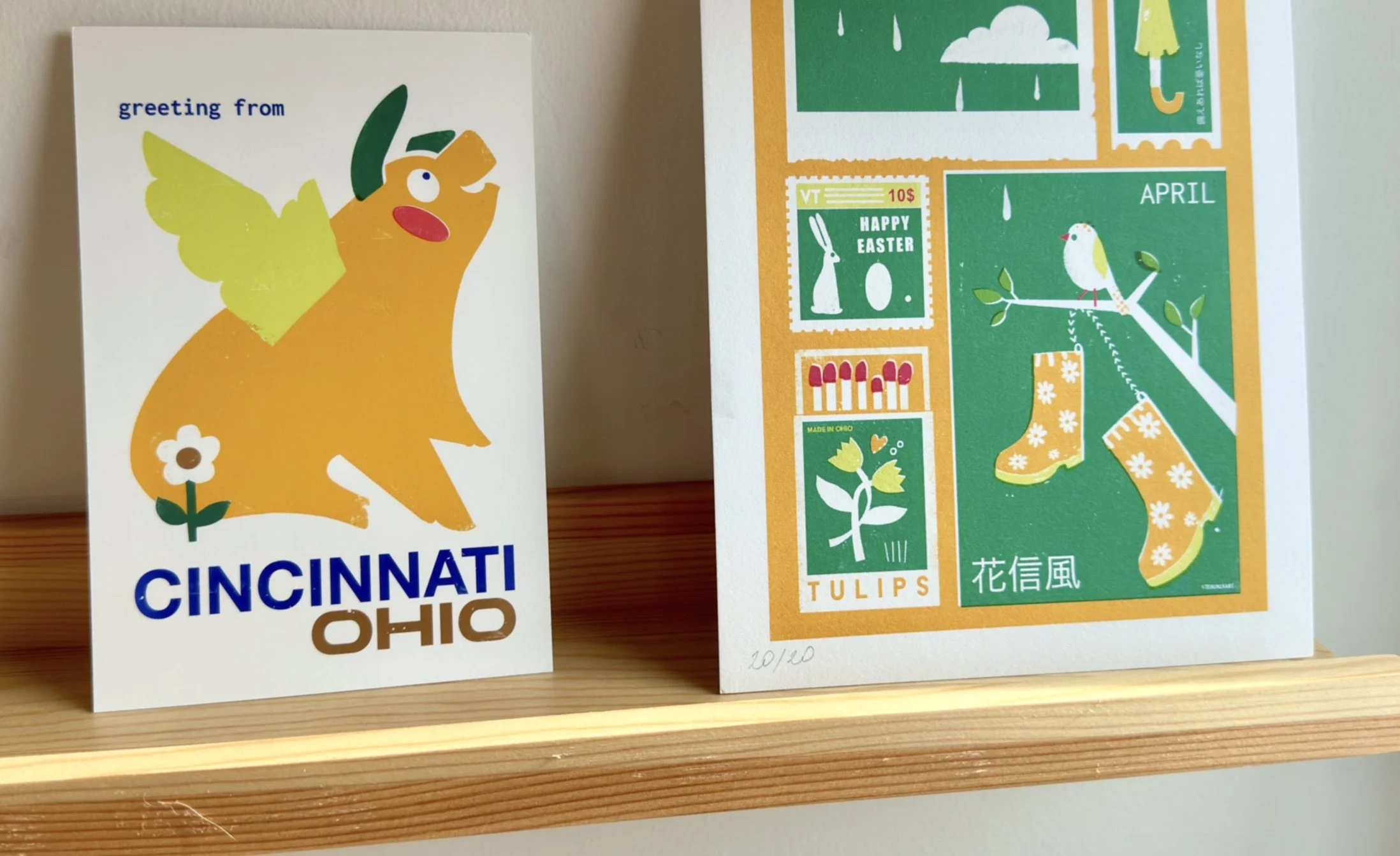 A Cincinnati Flying Pig postcard and a green and orange April illustration print displayed standing on a wooden shelf against a neutral wall.
