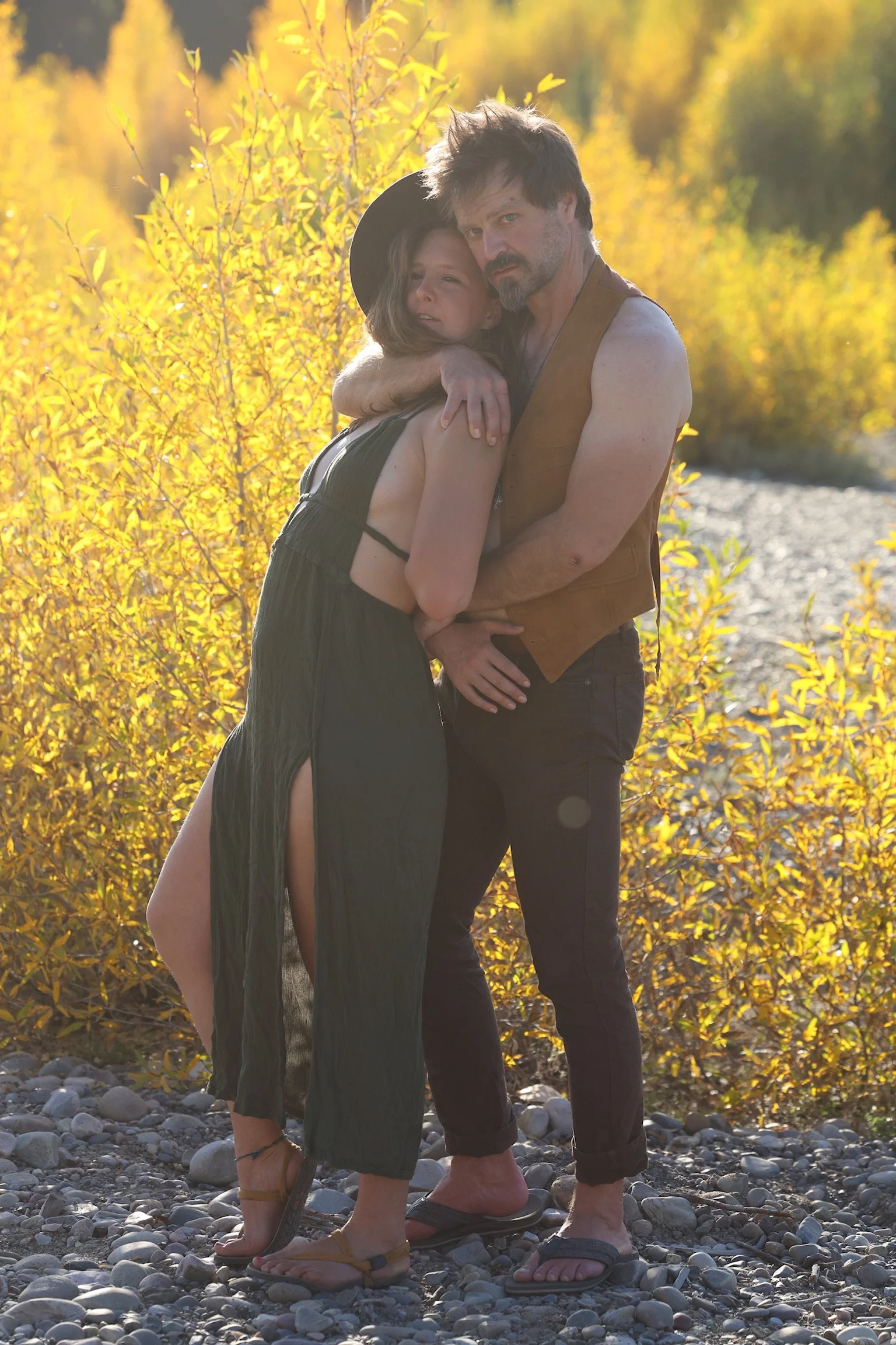 Two people hugging on a rocky riverbank during autumn with yellow foliage in the background.
