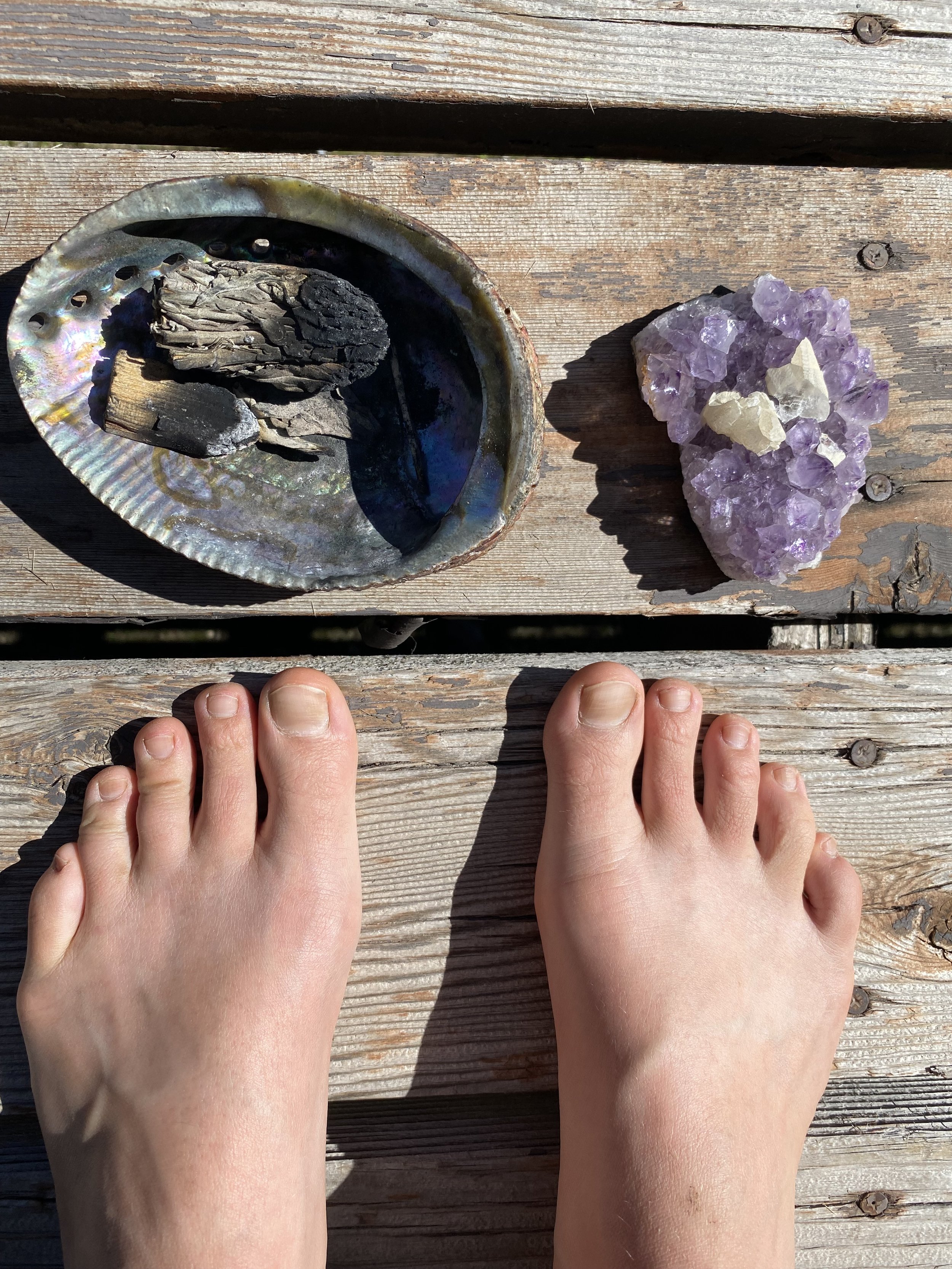 Two hands with light skin resting on a weathered wooden surface divided into sections by nails, with one shell containing blackened, charred wood pieces and the other displaying purple amethyst crystals.