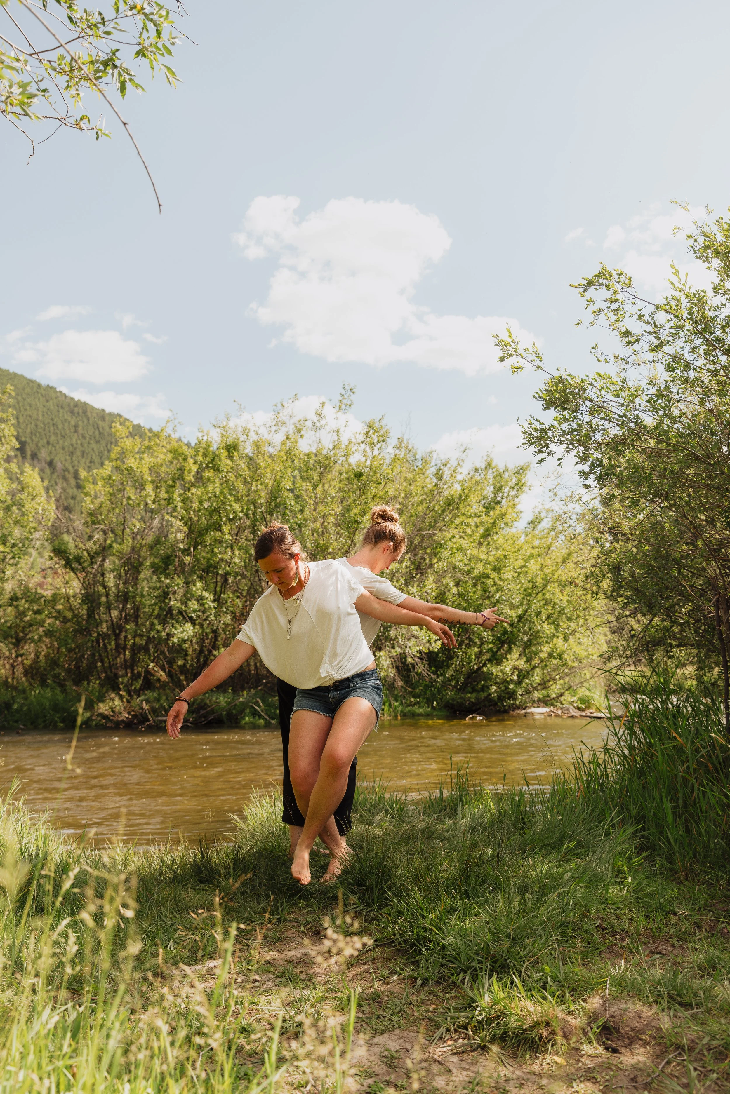 Two women with light skin are walking in water near a grassy riverbank in a natural landscape with green trees and blue sky with clouds.