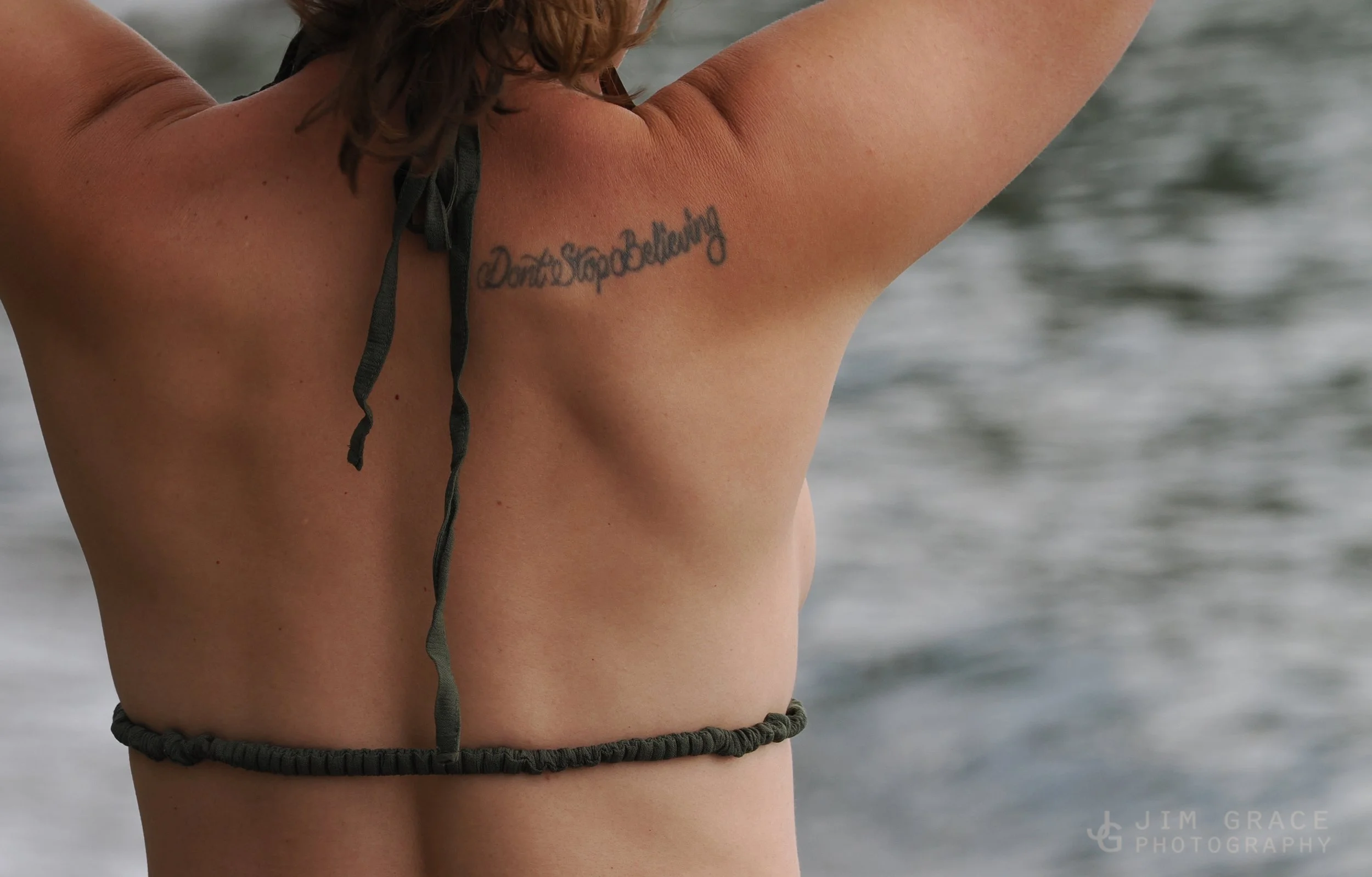 A woman with a tattoo on her upper chest near her shoulder, standing on a beach with the ocean in the background.
