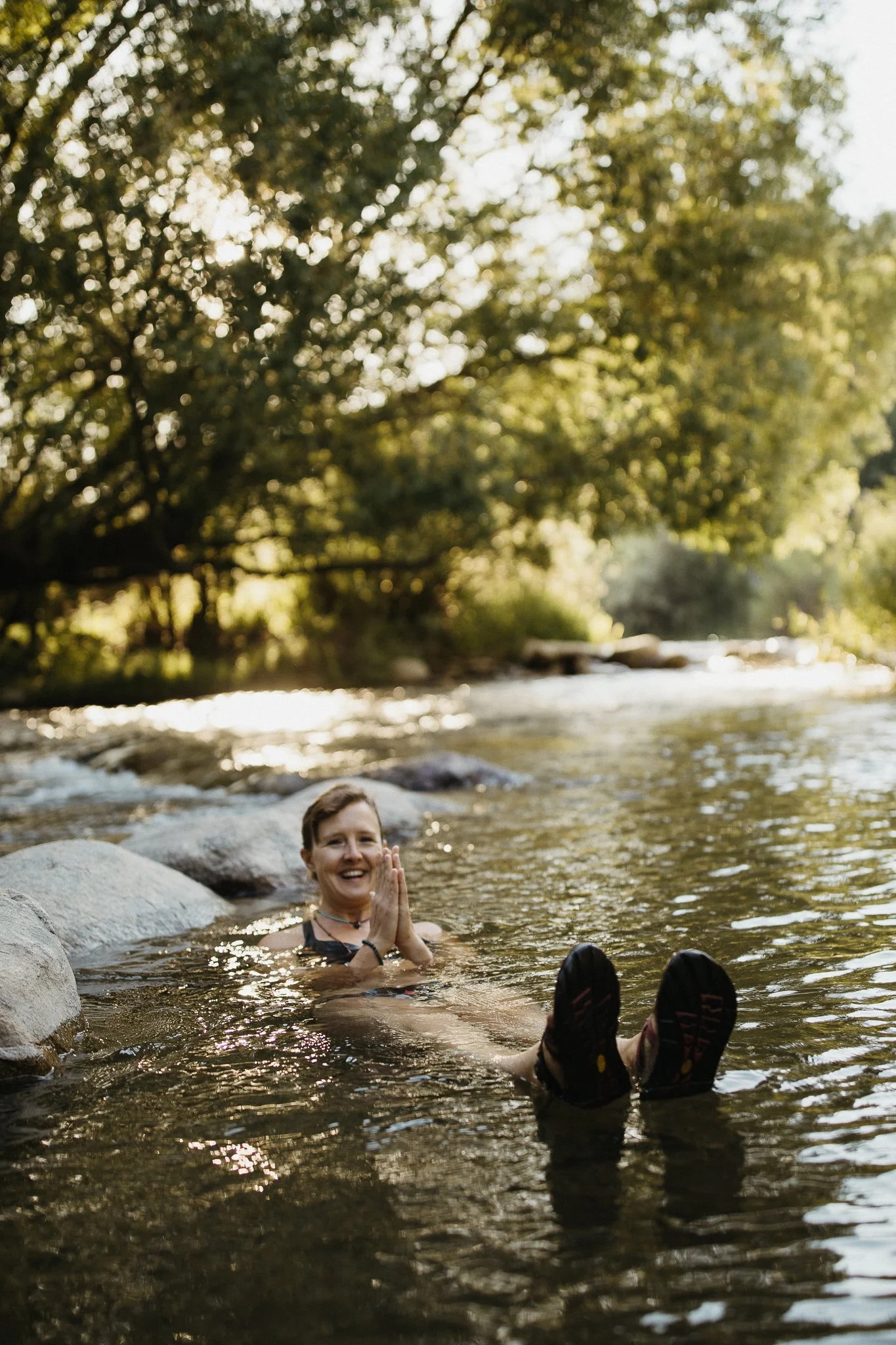Woman with short hair and a black top relaxing in a river, hands clasped, smiling, with legs stretched out and rocks around, trees with green leaves in the background and sunlight filtering through.