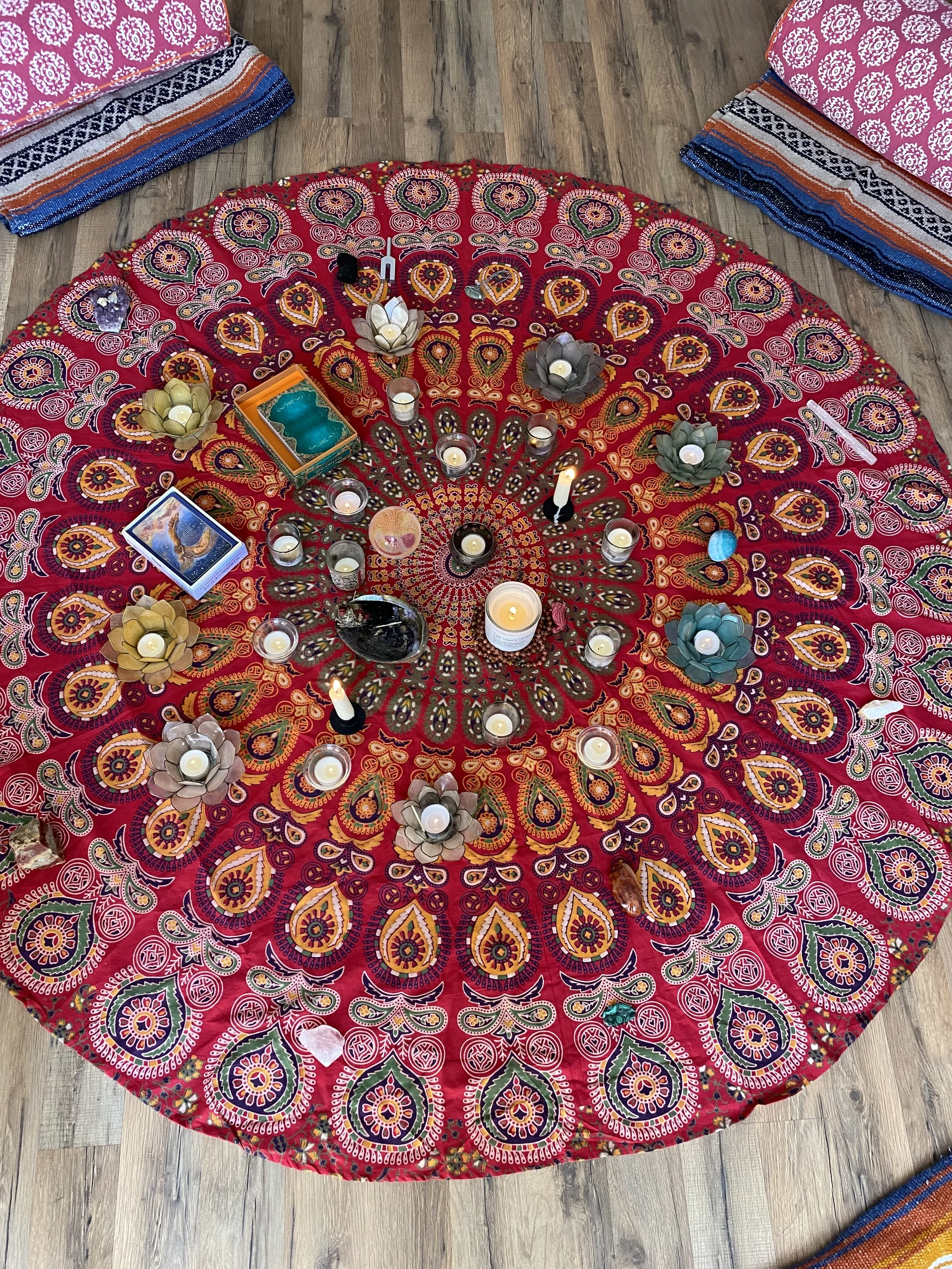 A circular table covered with a vibrant red tapestry featuring intricate floral and paisley patterns. The table has small candles, flower-shaped candle holders, a deck of tarot or oracle cards, and various stones or crystals arranged on it. Surrounding the table are textile cushions or mats with colorful, patterned designs, placed on a wooden floor.