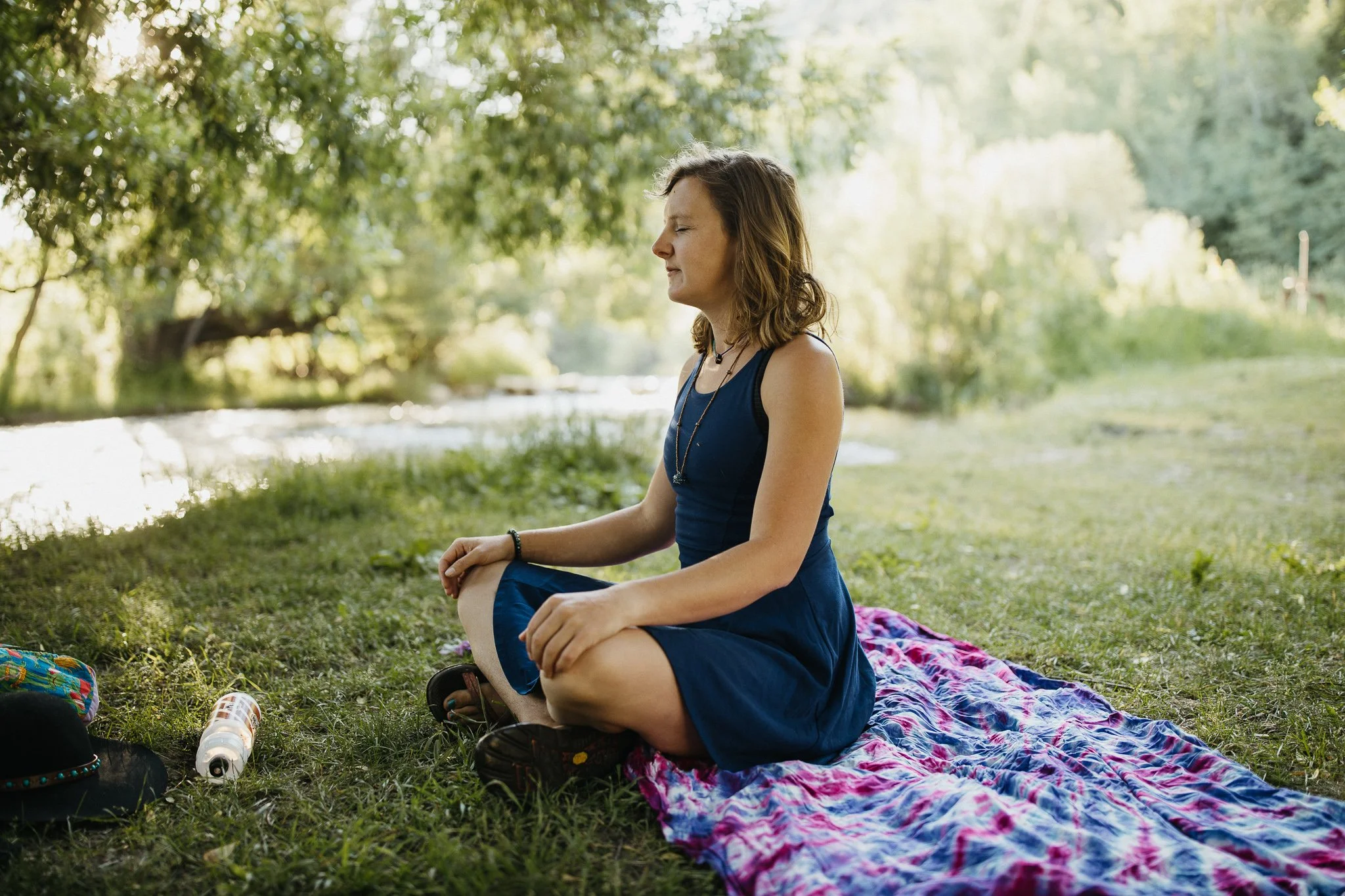 A woman practicing meditation outdoors by a river, sitting cross-legged on a colorful blanket with closed eyes and a peaceful expression.