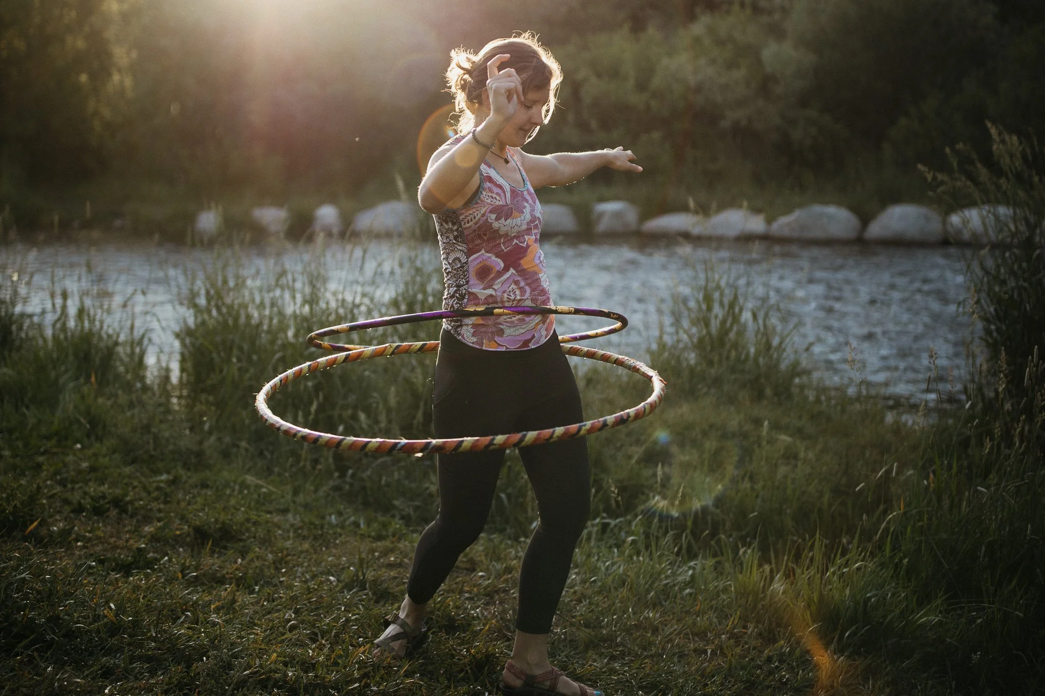 A woman in a floral tank top and black leggings hula hooping outdoors near a river at sunset.