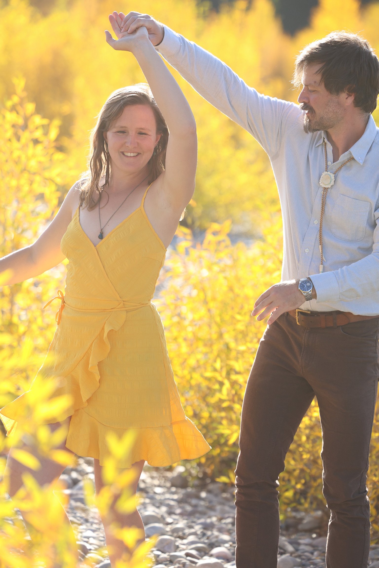A couple dancing outdoors in a park during autumn, with the woman in a yellow dress and the man in a light shirt and dark pants, surrounded by yellow foliage.
