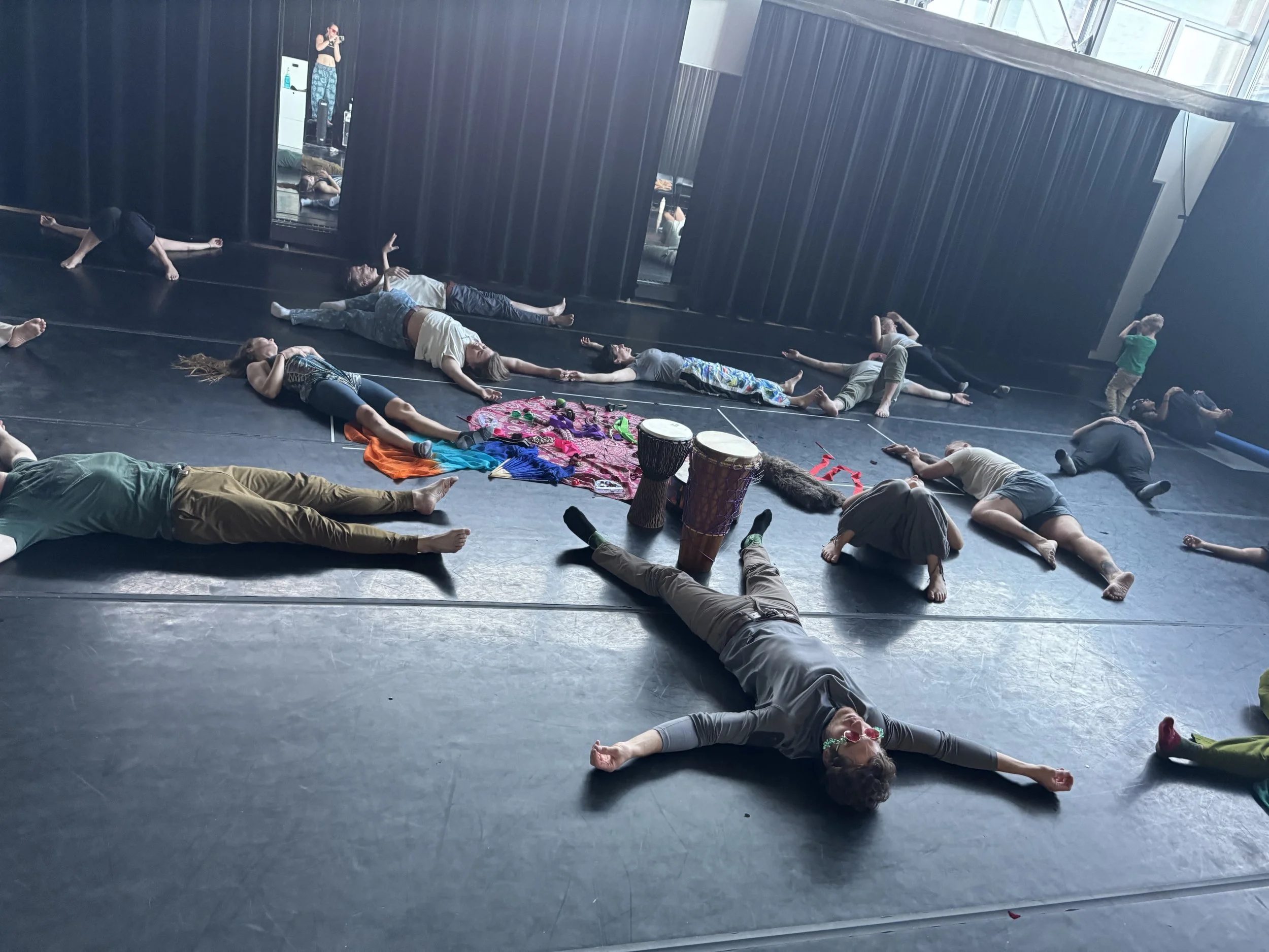 People lying on the floor of a dance studio, some holding hands, with various colorful props and musical drums around them, and a person with sunglasses lying in the foreground.
