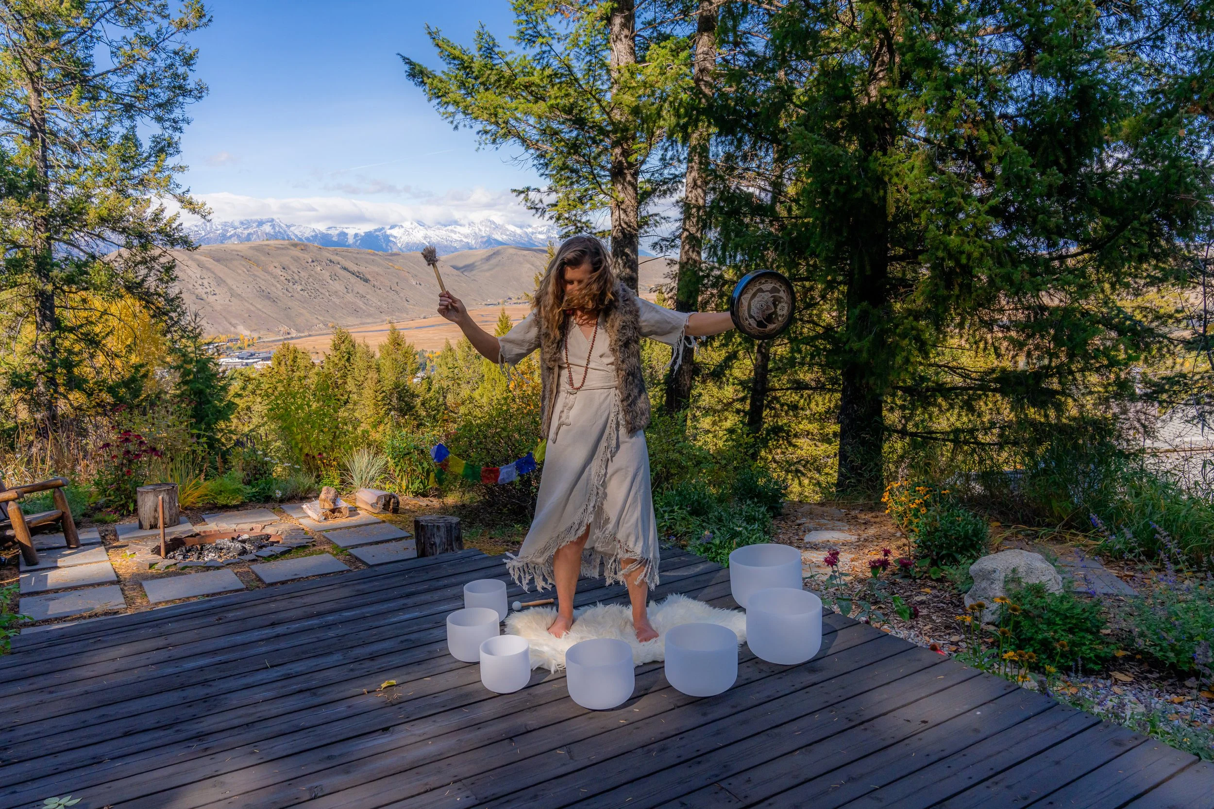 A woman dressed in bohemian style standing on a wooden deck outdoors, surrounded by trees and flowers with mountains in the background. She is holding a drum and a wand, with large candles around her, suggesting a spiritual or ritual setting.