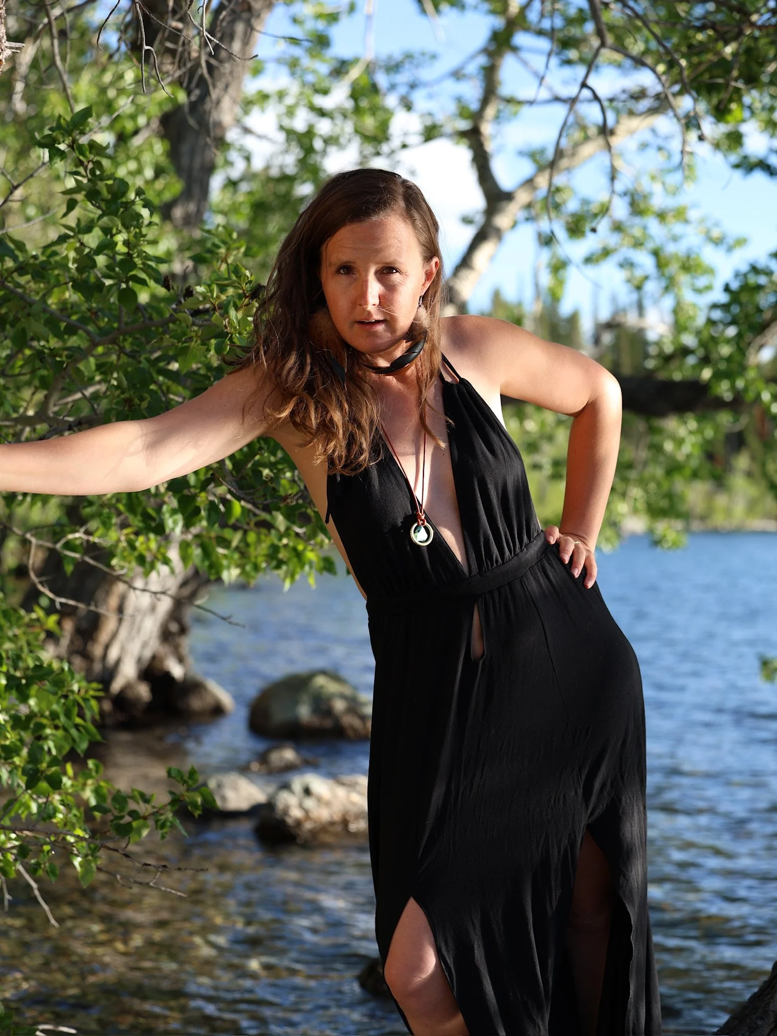 Woman in a black dress posing outdoors near a body of water and trees with green leaves, under a blue sky.