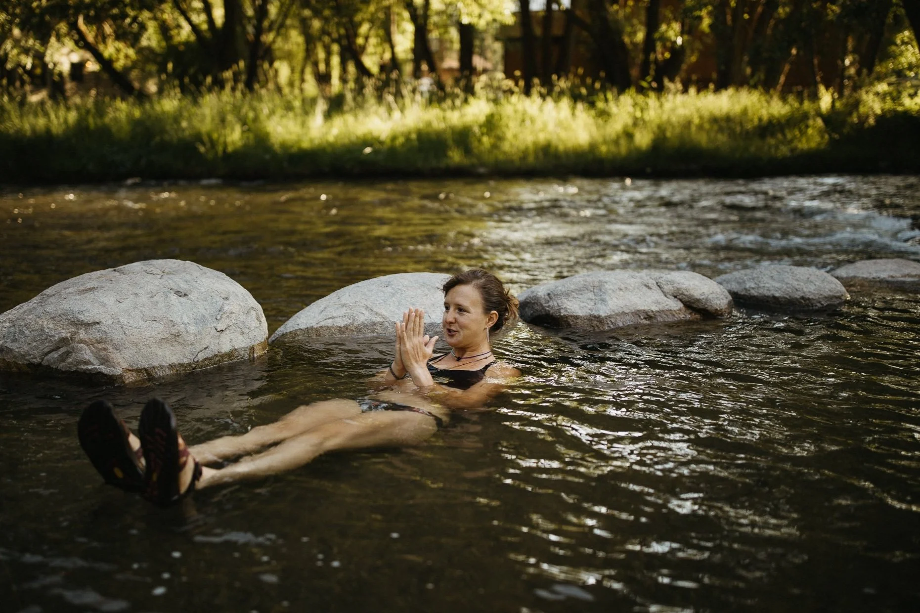 A woman with short dark hair, wearing a black swimsuit, relaxing in a shallow river with rocks around her. She is lying on her back with her legs stretched out and her hands in a prayer position. The background shows a lush, green, sunlit forest.