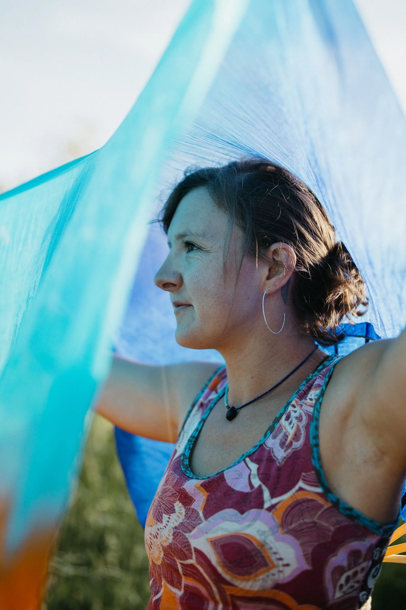 A woman with short hair, wearing a colorful patterned top, earrings, and a necklace, stands outdoors under a sheer blue fabric. The background is slightly blurred with greenery and a clear sky.