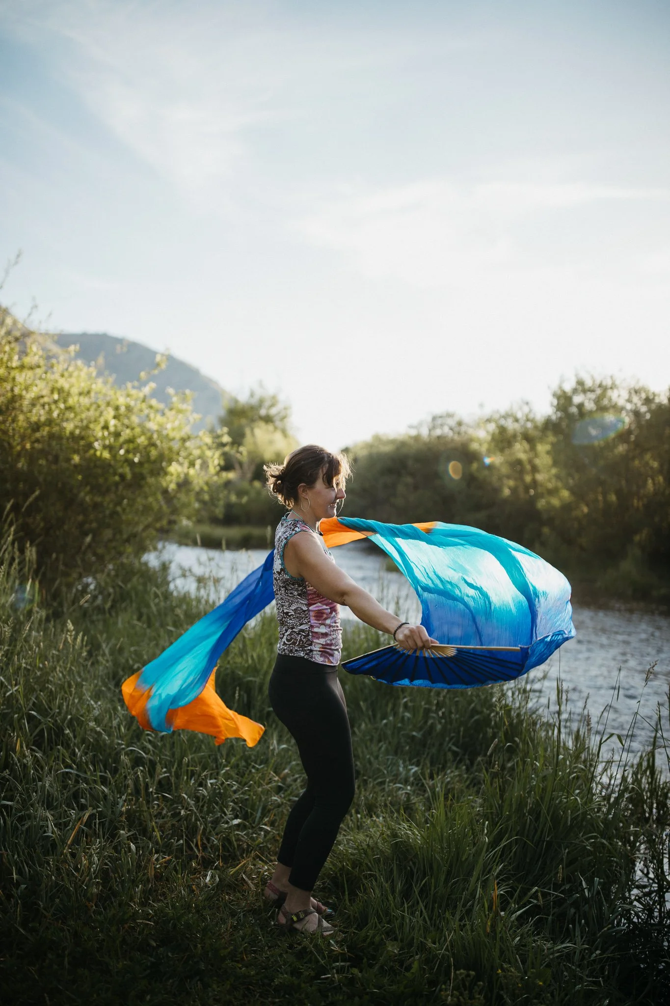 Woman standing on grassy riverbank holding colorful fabric banner, with trees, a river, mountains, and a clear sky in the background.