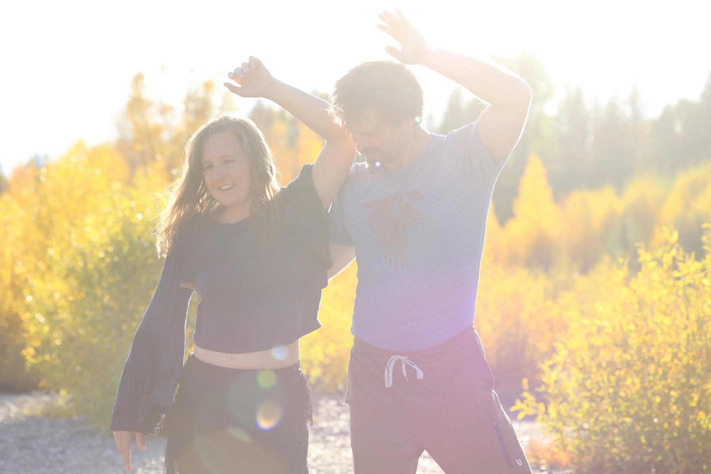 A young man and woman are standing outdoors in a sunny, fall environment, smiling, with trees showing autumn colors in the background.