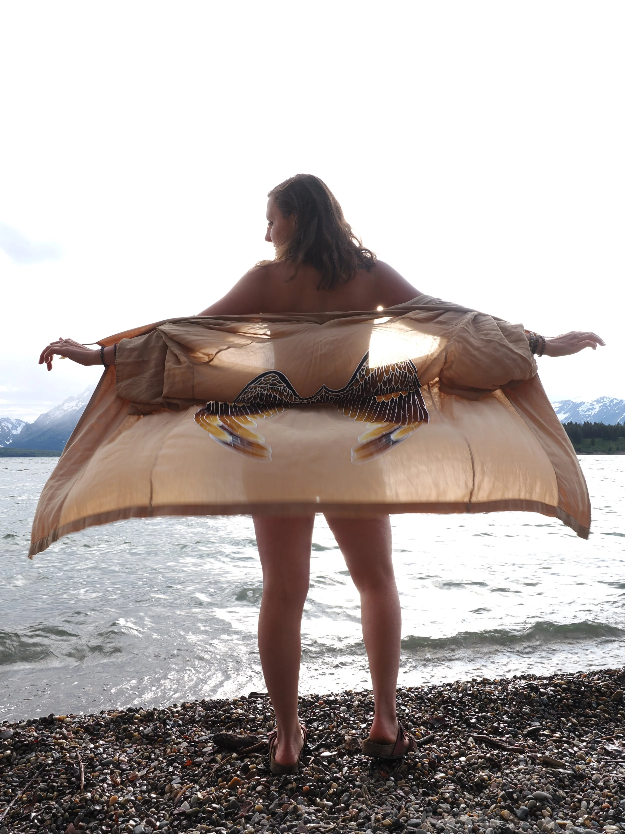 A woman standing on a pebble beach holding a large painted turtle shell, facing a body of water with mountains in the background, dressed in casual clothing.