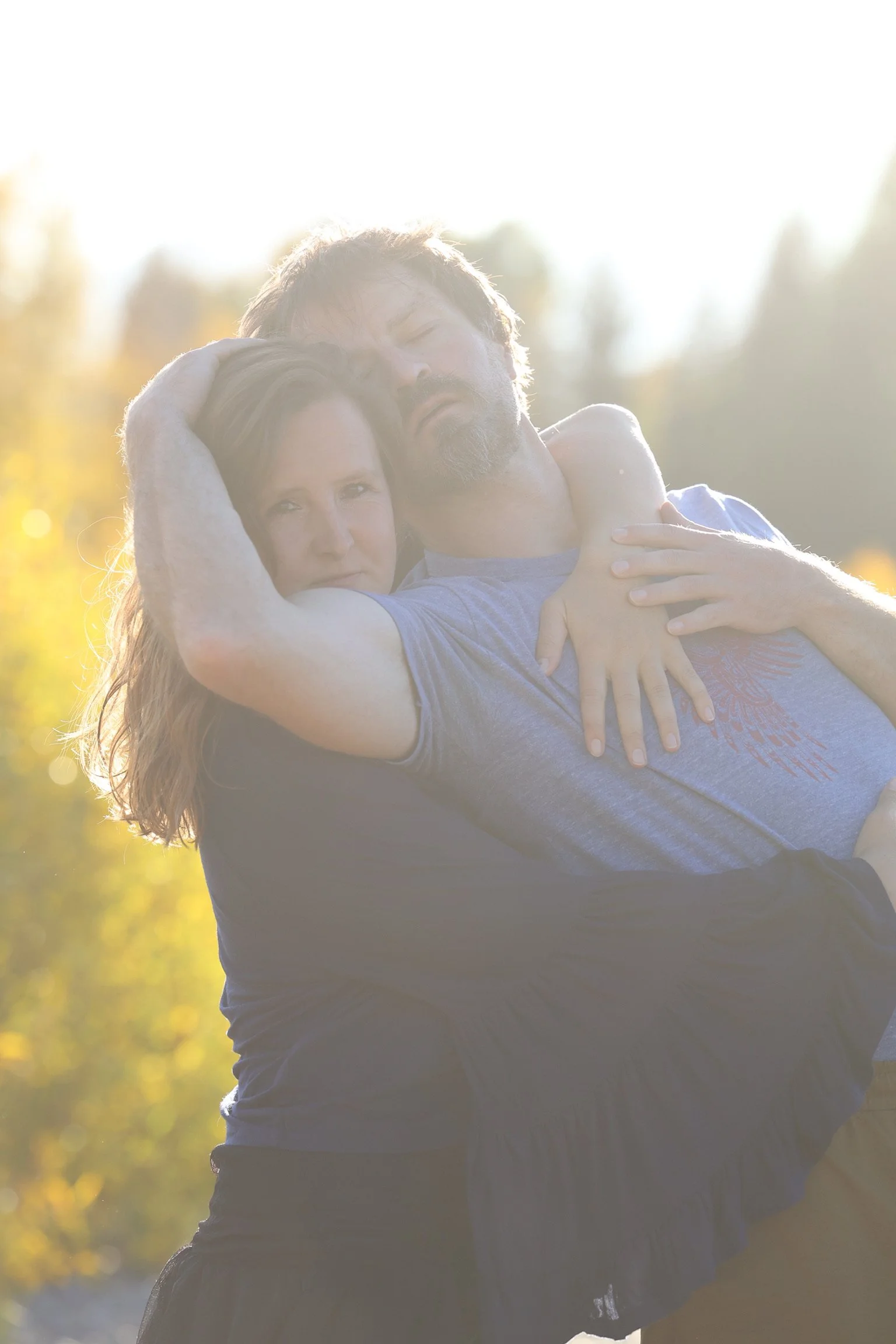 A couple embracing outdoors during sunset, with a bright, glowing sky and blurred trees in the background.