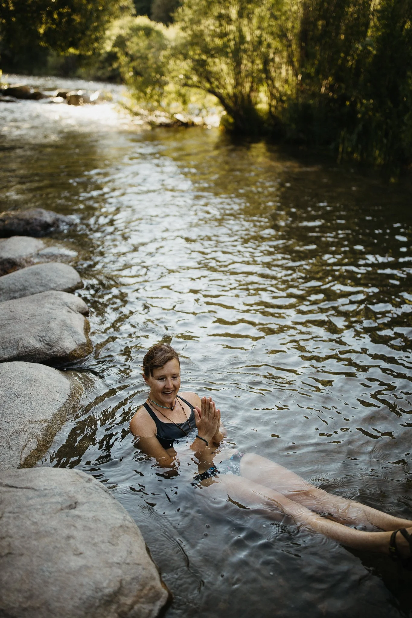 Woman practicing yoga in river, sitting on rocks, smiling, surrounded by trees