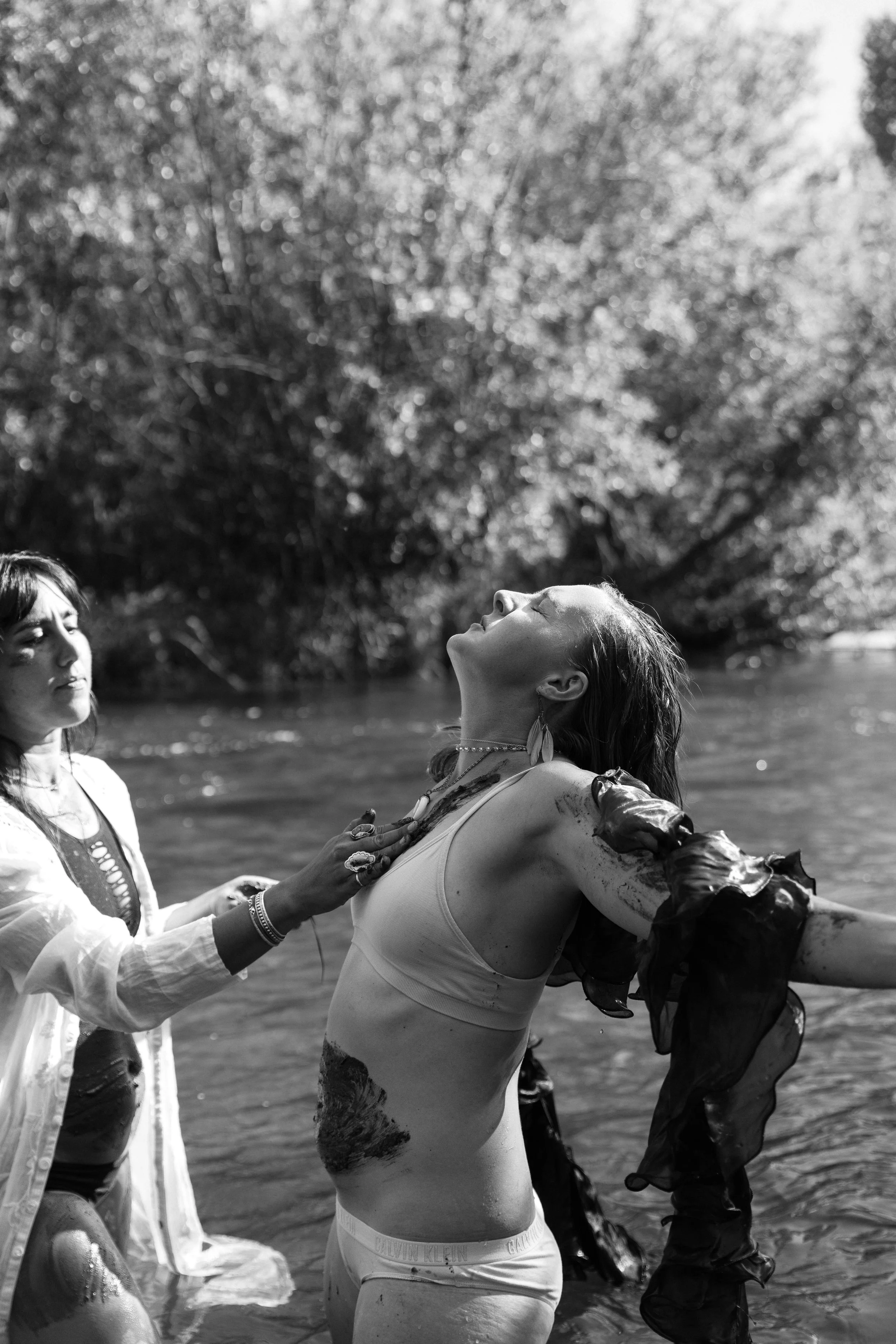 A woman with closed eyes and a serene expression being gently supported by another woman in a flowing dress near a body of water, with trees in the background, in a black-and-white photograph.