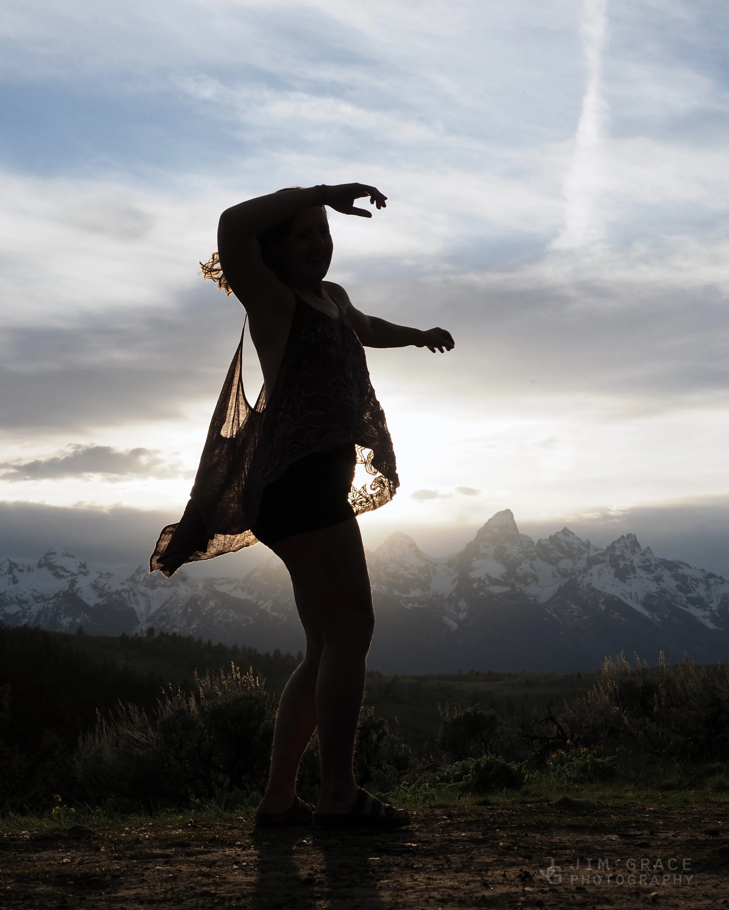 Silhouette of a woman standing outdoors against a mountainous landscape, with clouds and a partly cloudy sky in the background.
