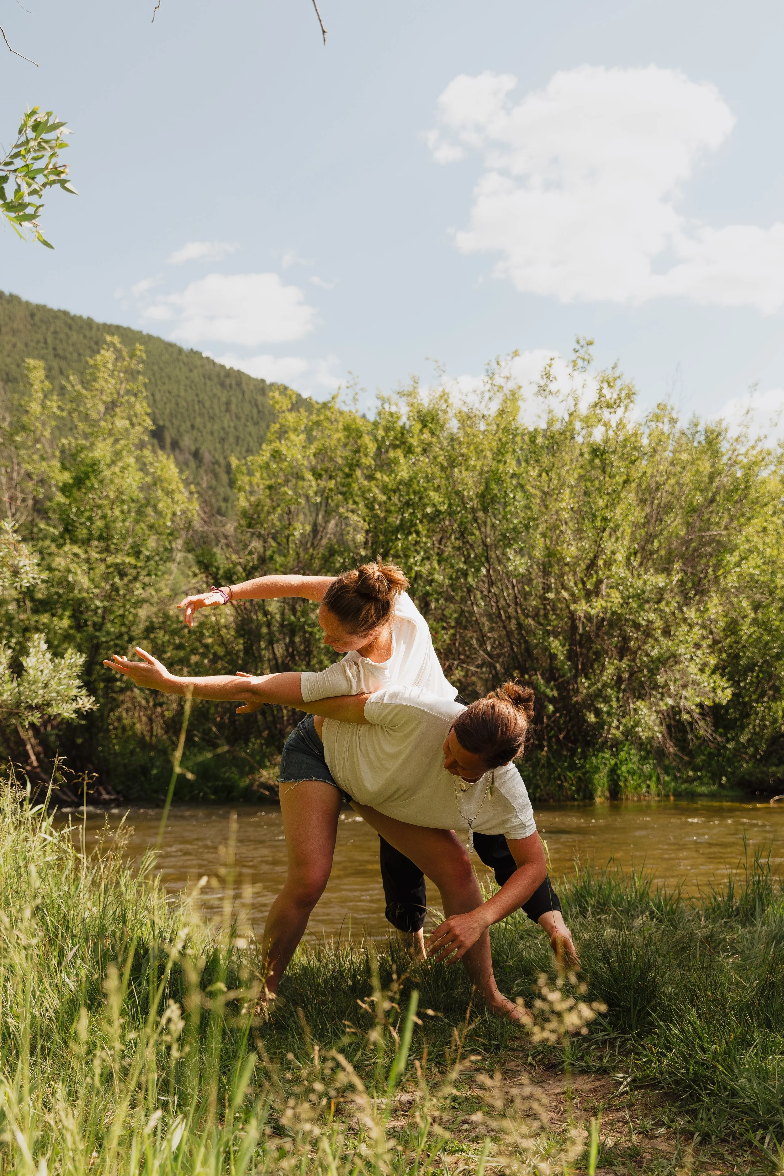 Two women performing an acro yoga pose by a river, surrounded by trees and greenery, with a mountain and blue sky with clouds in the background.