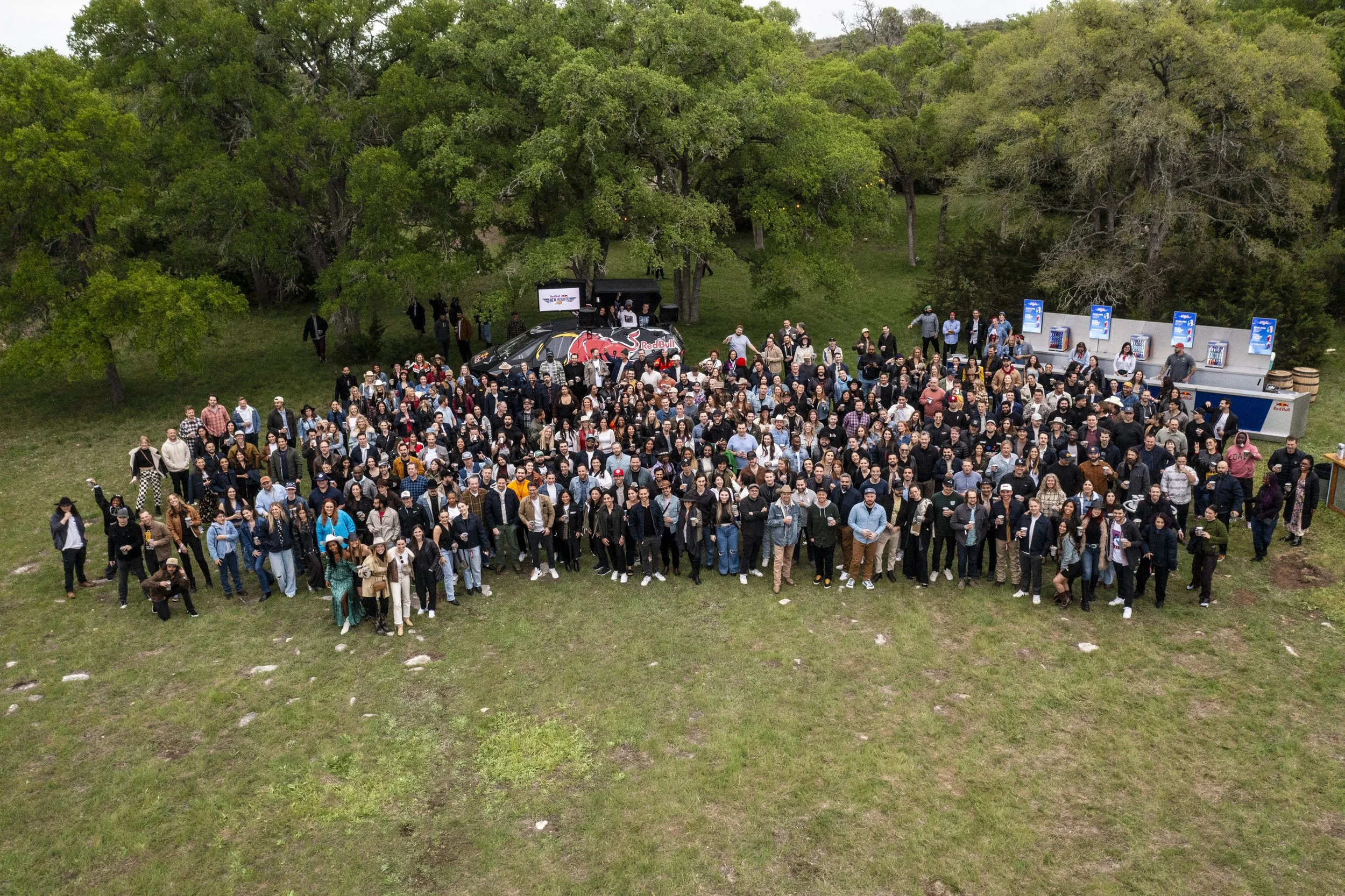 A large crowd of people gathered outdoors on a grassy field surrounded by trees, with a Red Bull racing vehicle and a food or drink stand in the background.
