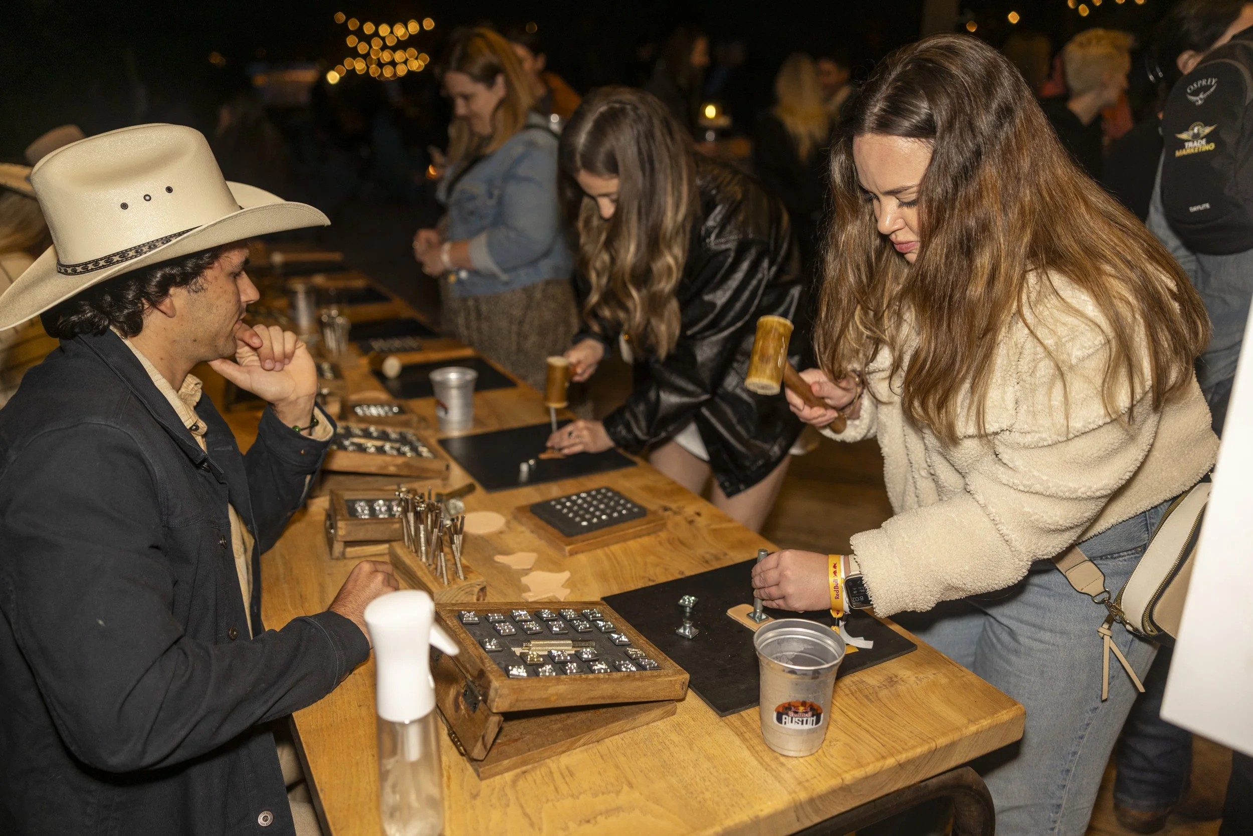 People playing a board game with pieces and cards at a wooden table, with one woman holding a hammer and a man in a cowboy hat watching.