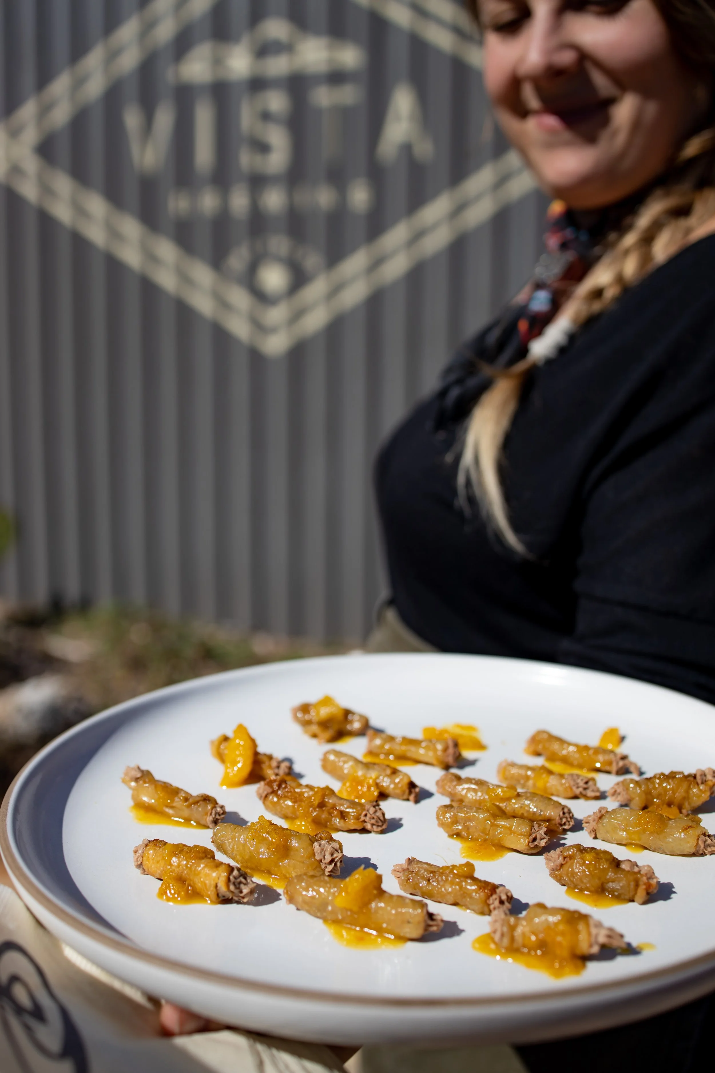 A person holding a white plate with small, fried appetizers topped with yellow sauce. The person's face is partially visible, and they are smiling. In the background, there is a gray wall with an indistinct logo or signage.