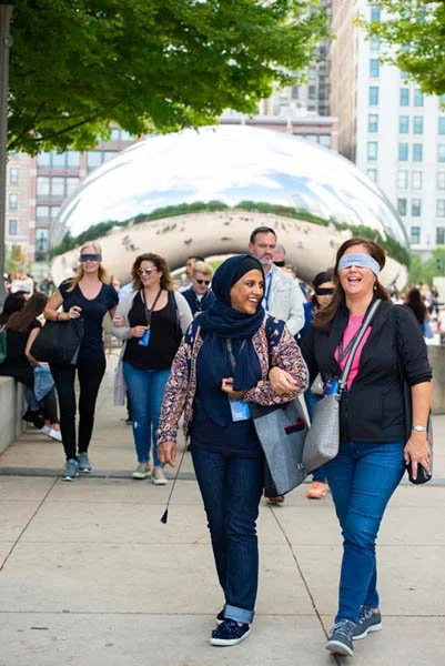 People walking outdoors near the Cloud Gate sculpture in Millennium Park, Chicago, with some wearing eye masks or glasses.