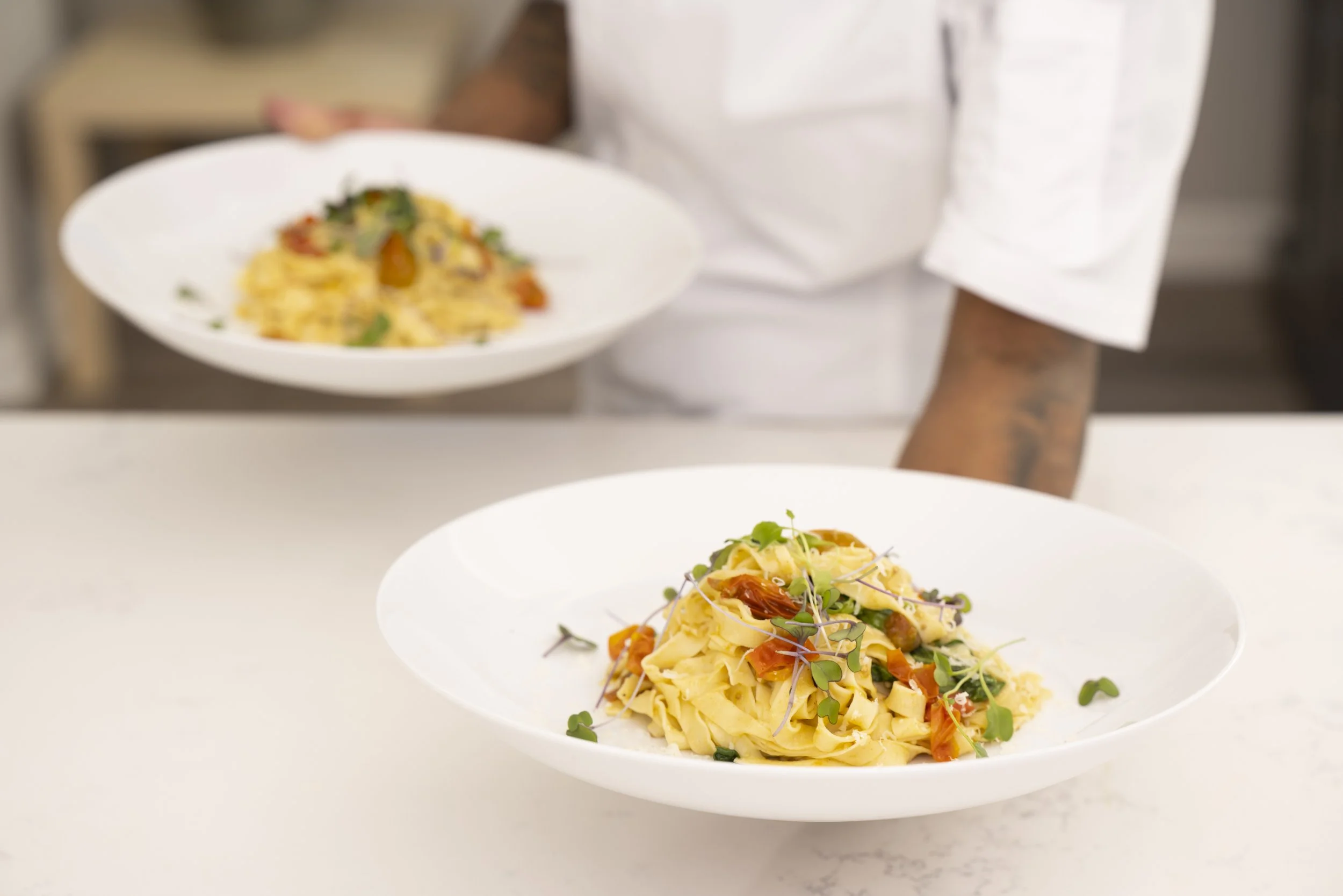 A chef in a white uniform holding a plate of pasta with vegetables, served on a white plate, on a white countertop.