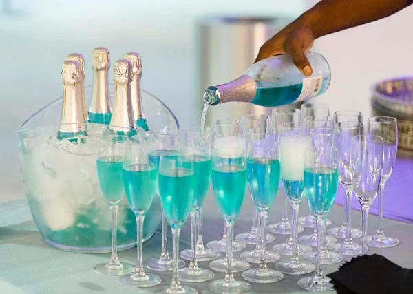 Person pouring blue cocktail into champagne flutes at a party, with bottles of champagne in an ice bucket nearby.