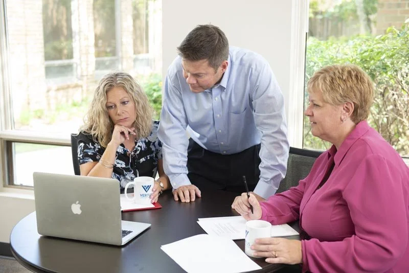 Three people having a discussion at a table with a laptop, papers, and coffee mugs in a bright room with large windows and greenery outside.