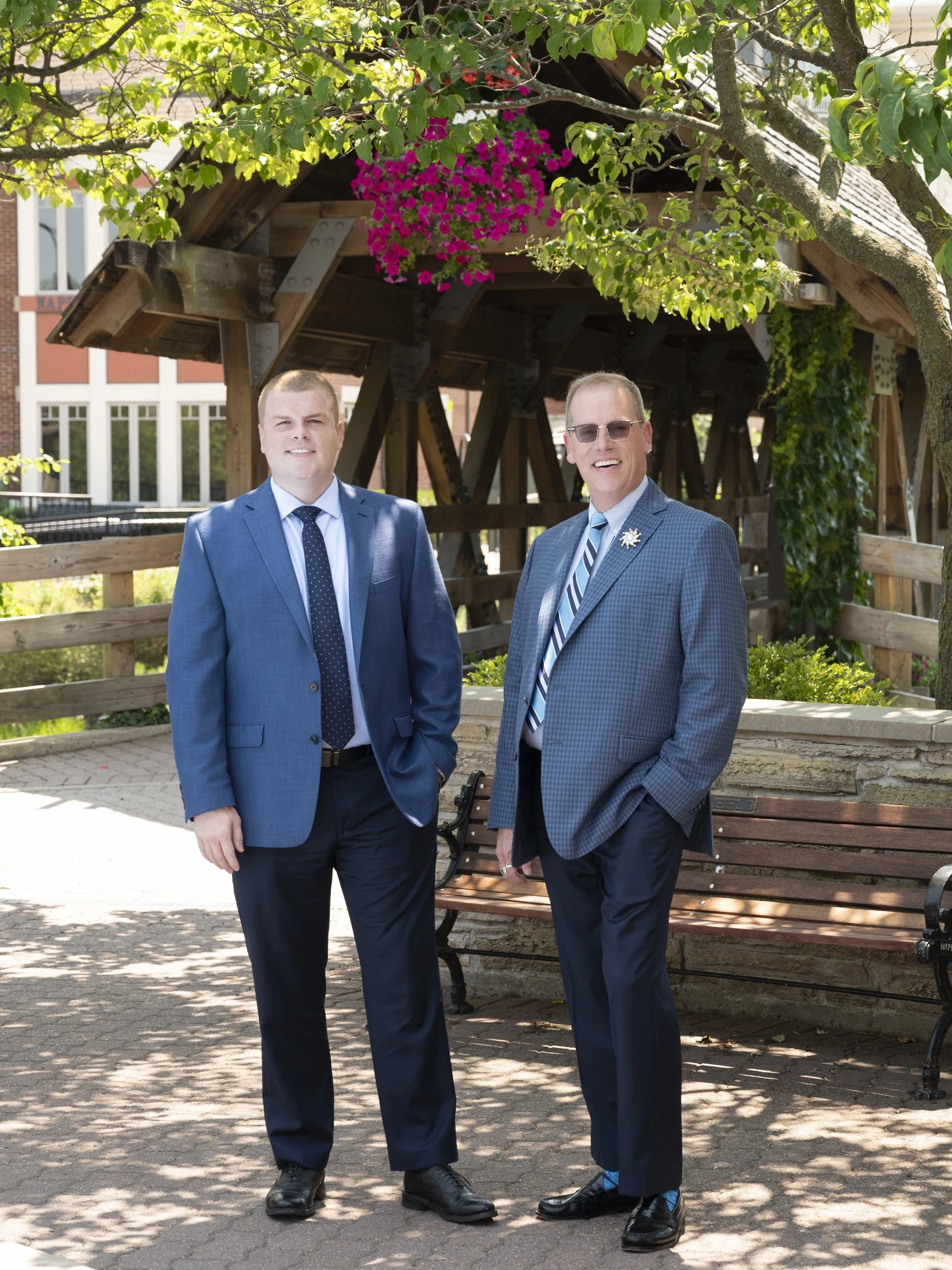 Group portrait photograpy of two real estate agents standing outdoors under a tree with pink flowers, with a wooden shelter behind them and benches nearby.