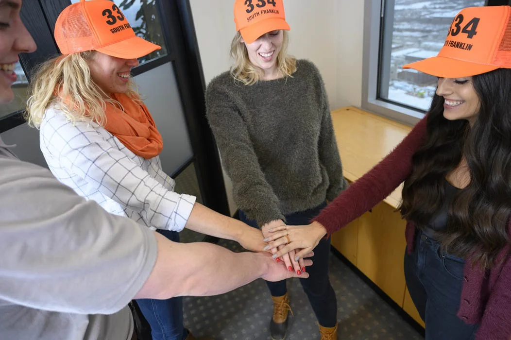 Four colleagues, three women and one man, wearing orange hats, place their hands together in a show of teamwork in an office with a window showing a cityscape during daytime.