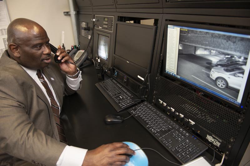 A man in a suit talking on a phone at a workstation with multiple computer monitors displaying security footage.