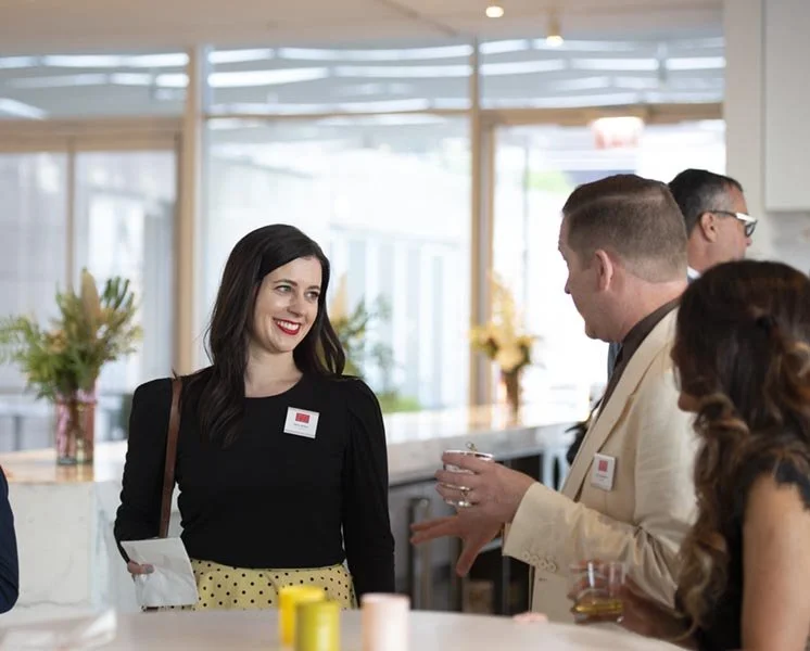 People talking at a social event in a bright room with large windows, flowers, and a counter.