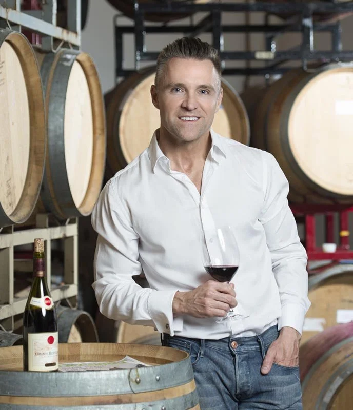 Chicago portrait photography of a sommelier inside a restaurant holding a glass of wine. He is in front of wine barrels with a bottle off the to left side of the frame. Only the best photographers can capture low light portraits in tricky environment