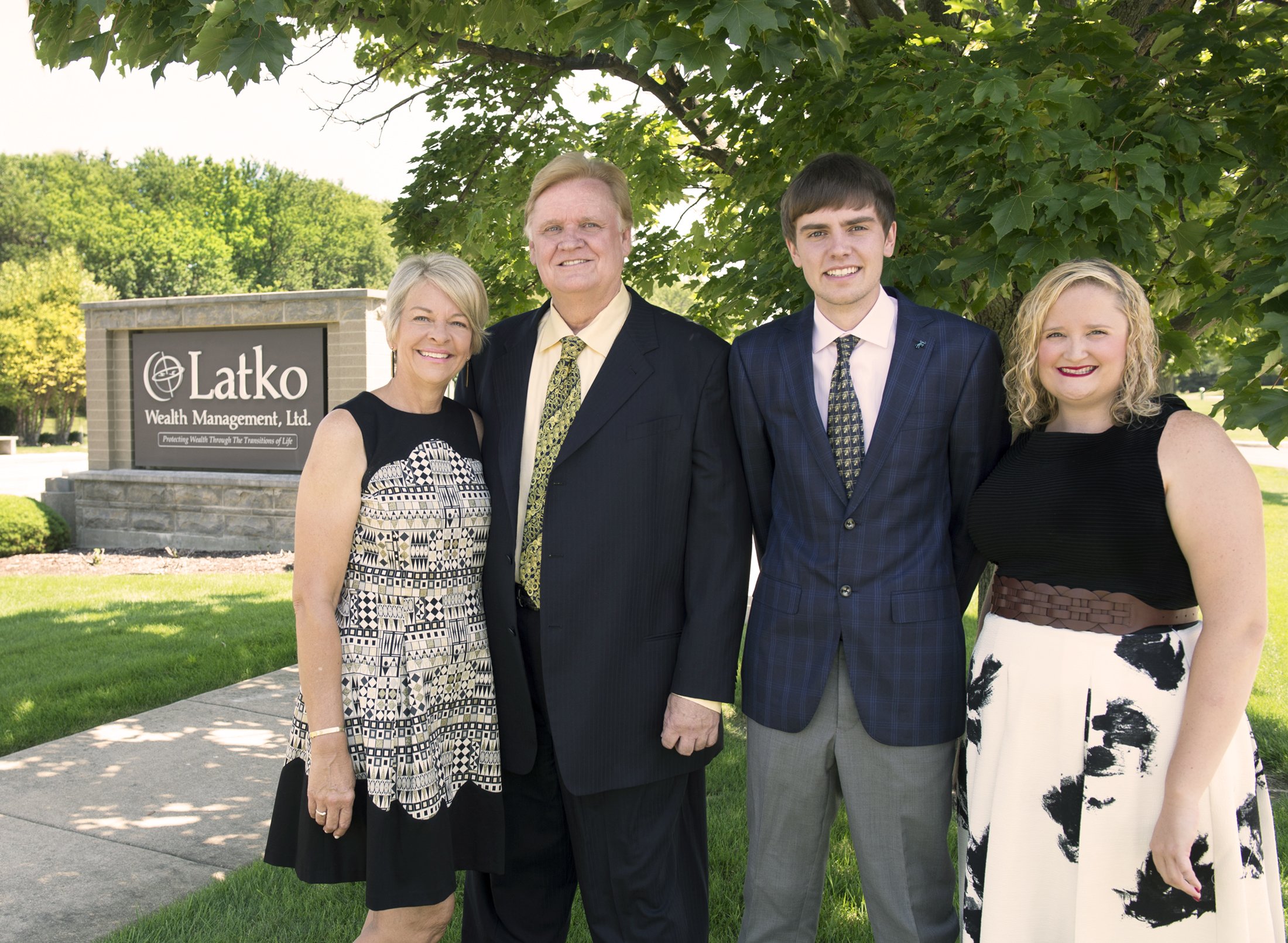 Four people standing outdoors in front of a sign that reads 'Latko Wealth Management Ltd.' with a tagline below. They are smiling, dressed in business or semi-formal attire, with trees and greenery in the background.