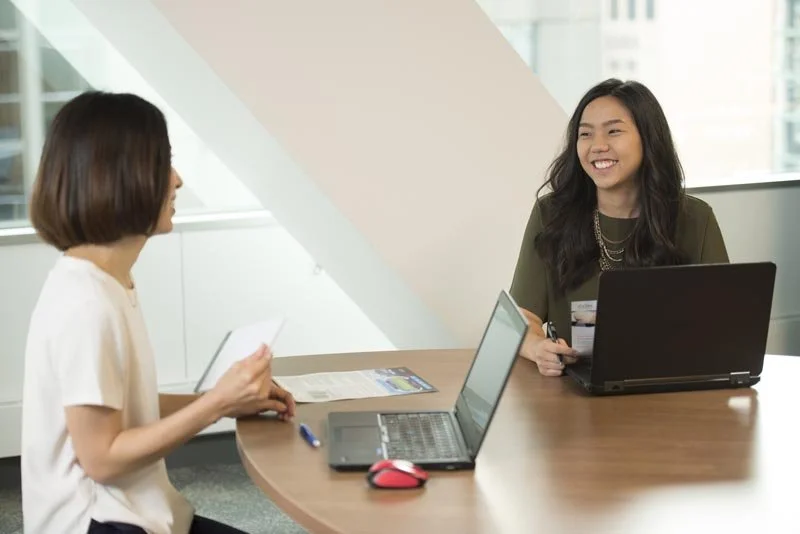 Two women smiling and having a discussion at a round wooden table with laptops in a modern office setting.