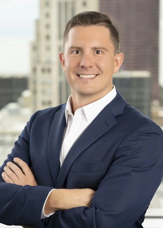 Executive portrait in a large law firm downtown in business attire and crossed arms. This  showcases a corporate brand by a top Chicago photographer taken with a blurred background in the company's office with large windows and a skyline view.