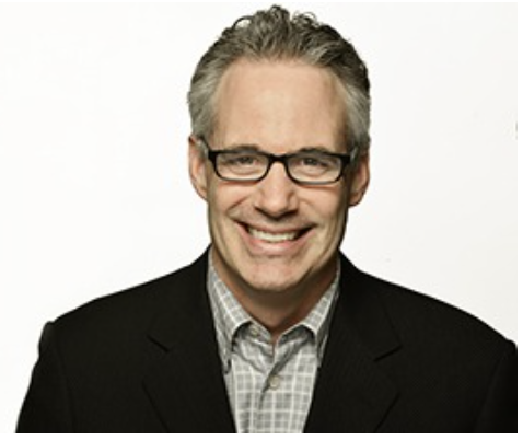 A headshot middle-aged man with gray hair, glasses, and a big smile, wearing a black blazer and a light-colored checkered shirt against a white background in Chicago.