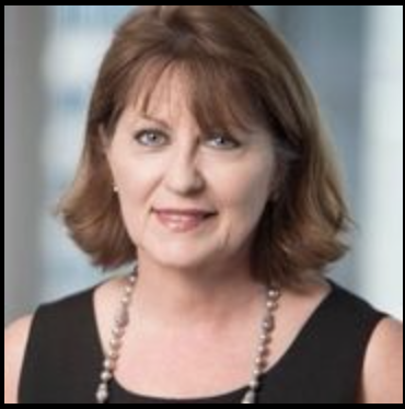 A woman with shoulder-length auburn hair, wearing a black top and a pearl necklace, smiling in an indoor setting.