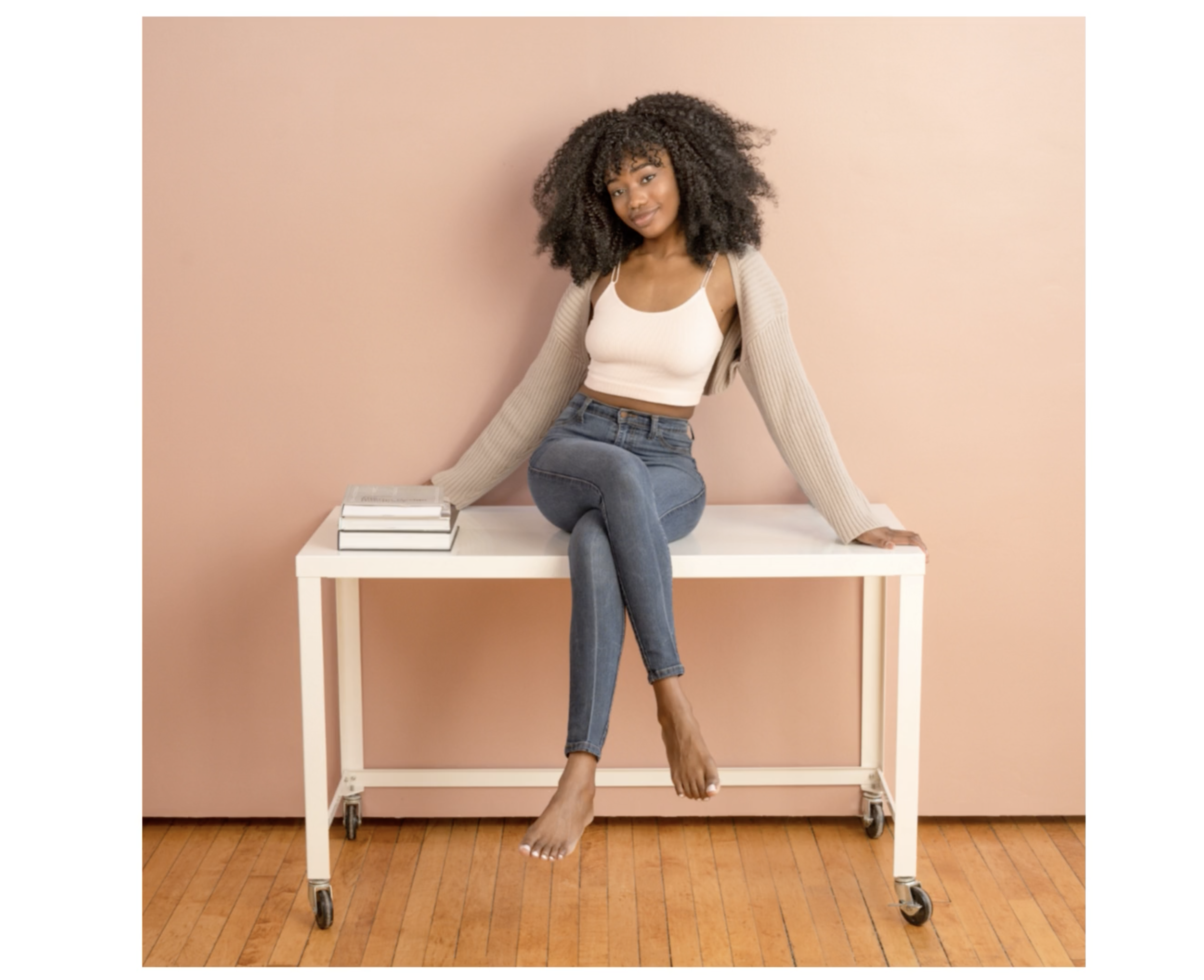 A social media influencer with curly hair sitting on a white table against a pink wall, wearing a beige cardigan, white tank top, and jeans, with a stack of books beside her for her personal branding marketing.
