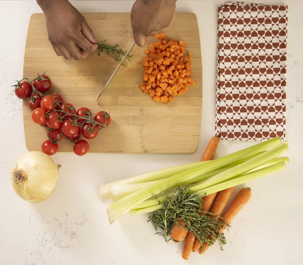 Person chopping carrots on a wooden cutting board with tomatoes, onion, celery, carrots, and herbs nearby.