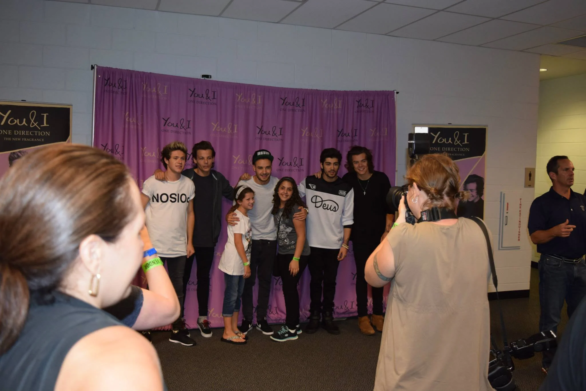 Group of six people, including young men, women, and two children, posing for a photo in front of a purple backdrop with 'You & I' and 'One Direction' written on it, while a woman with a camera takes their picture.