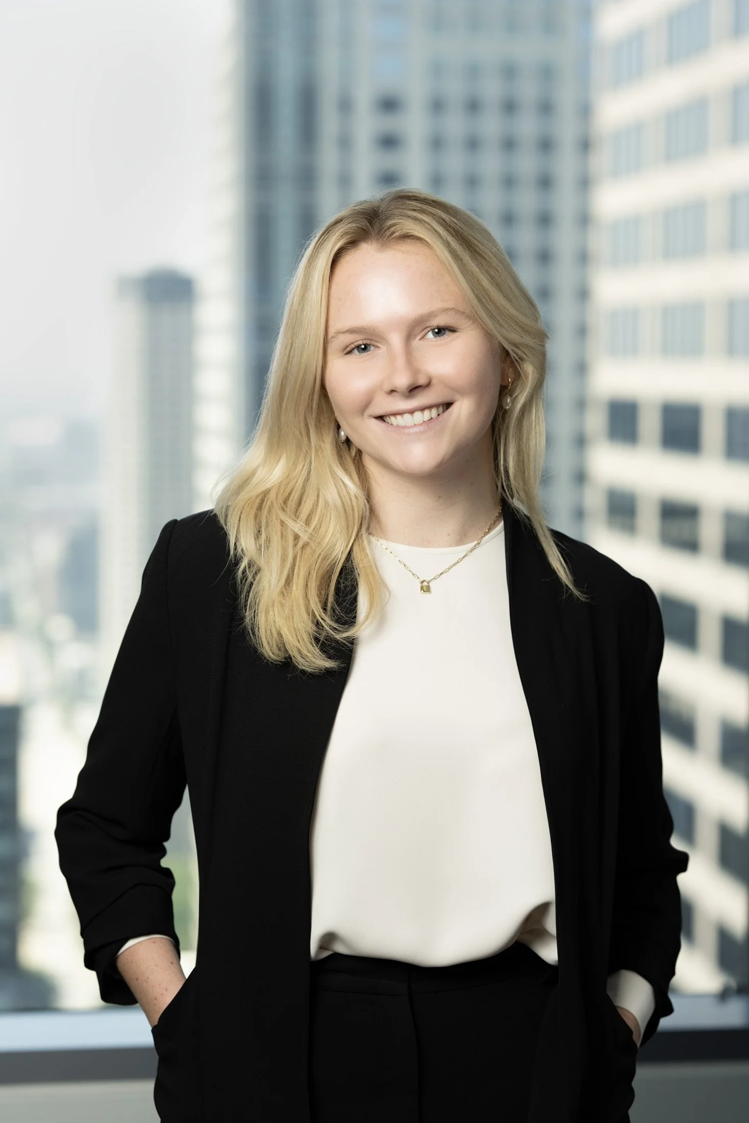 A young woman with blonde hair, wearing a black blazer and white blouse, smiling in front of a window with a cityscape background.