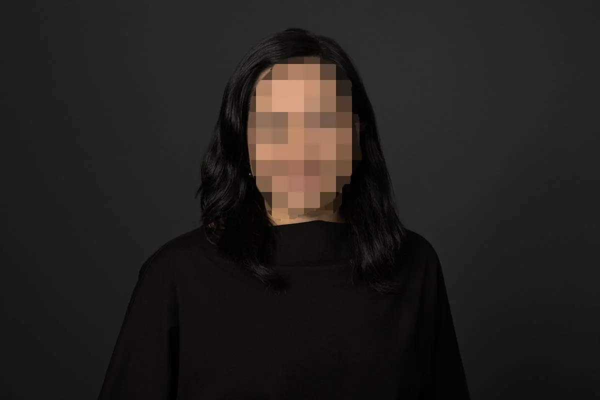 Portrait of a woman with black hair wearing a black top against a dark background, with her face pixelated to obscure her identity.