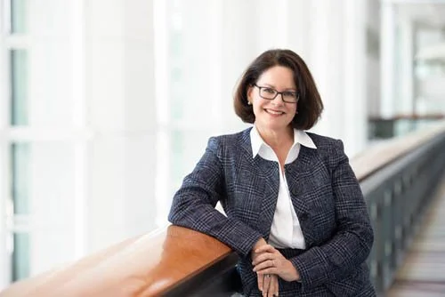 A smiling woman in professional attire leaning on a wooden railing in a bright, modern office building.